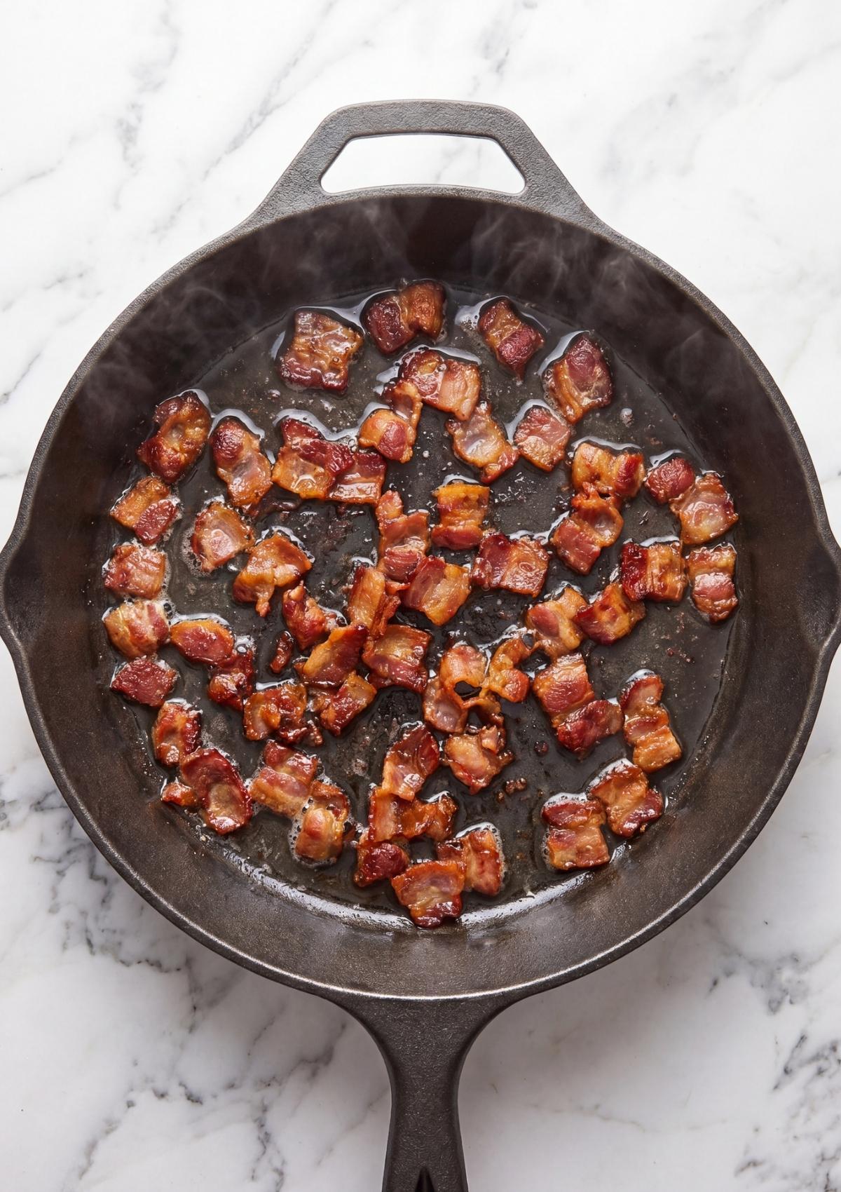 Small pieces of bacon frying until crispy in a dark cast iron skillet with visible steam.