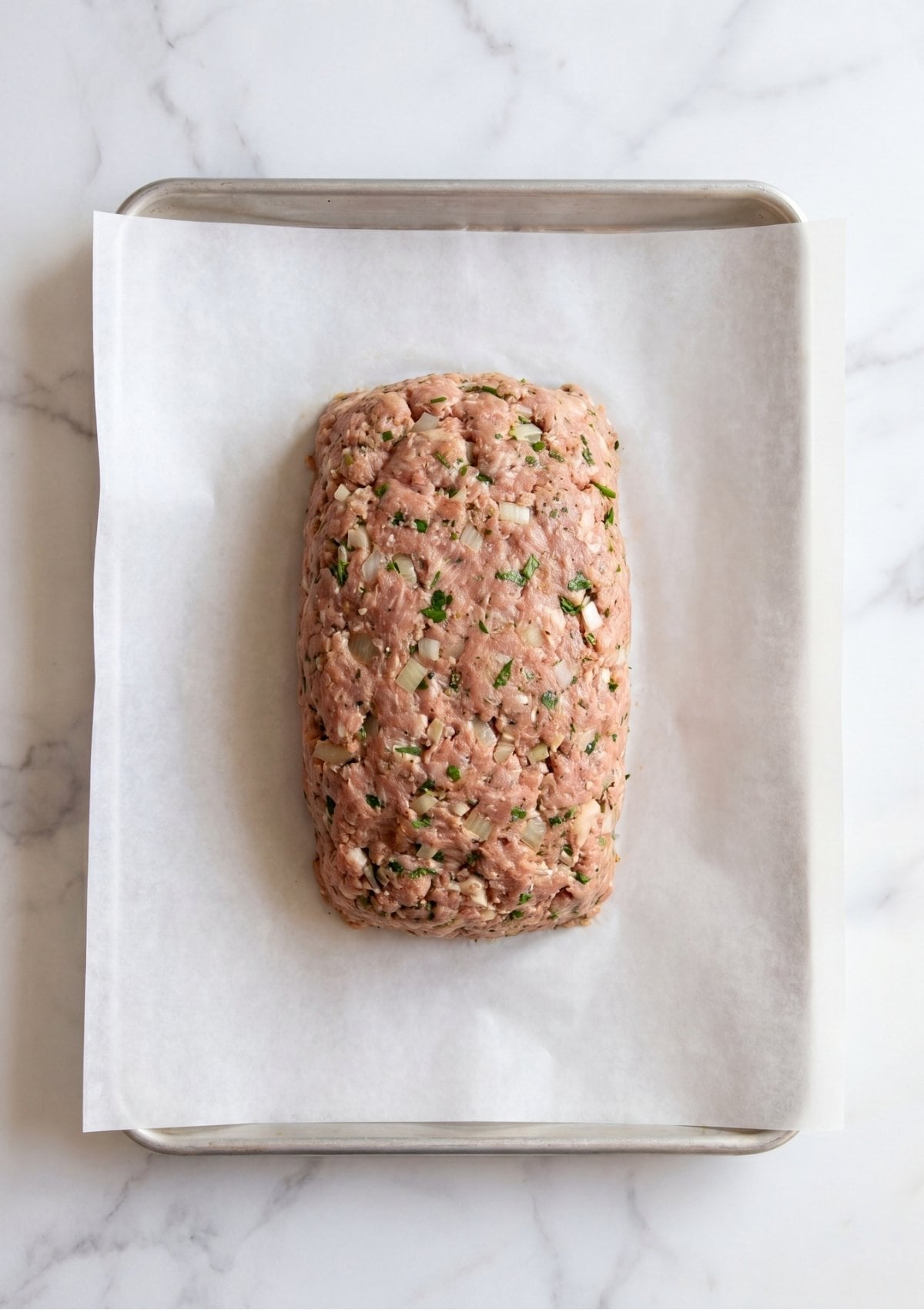 Overhead view of a raw, neatly shaped meatloaf mixture with visible onion pieces and green herbs sitting on a parchment paper lined baking sheet