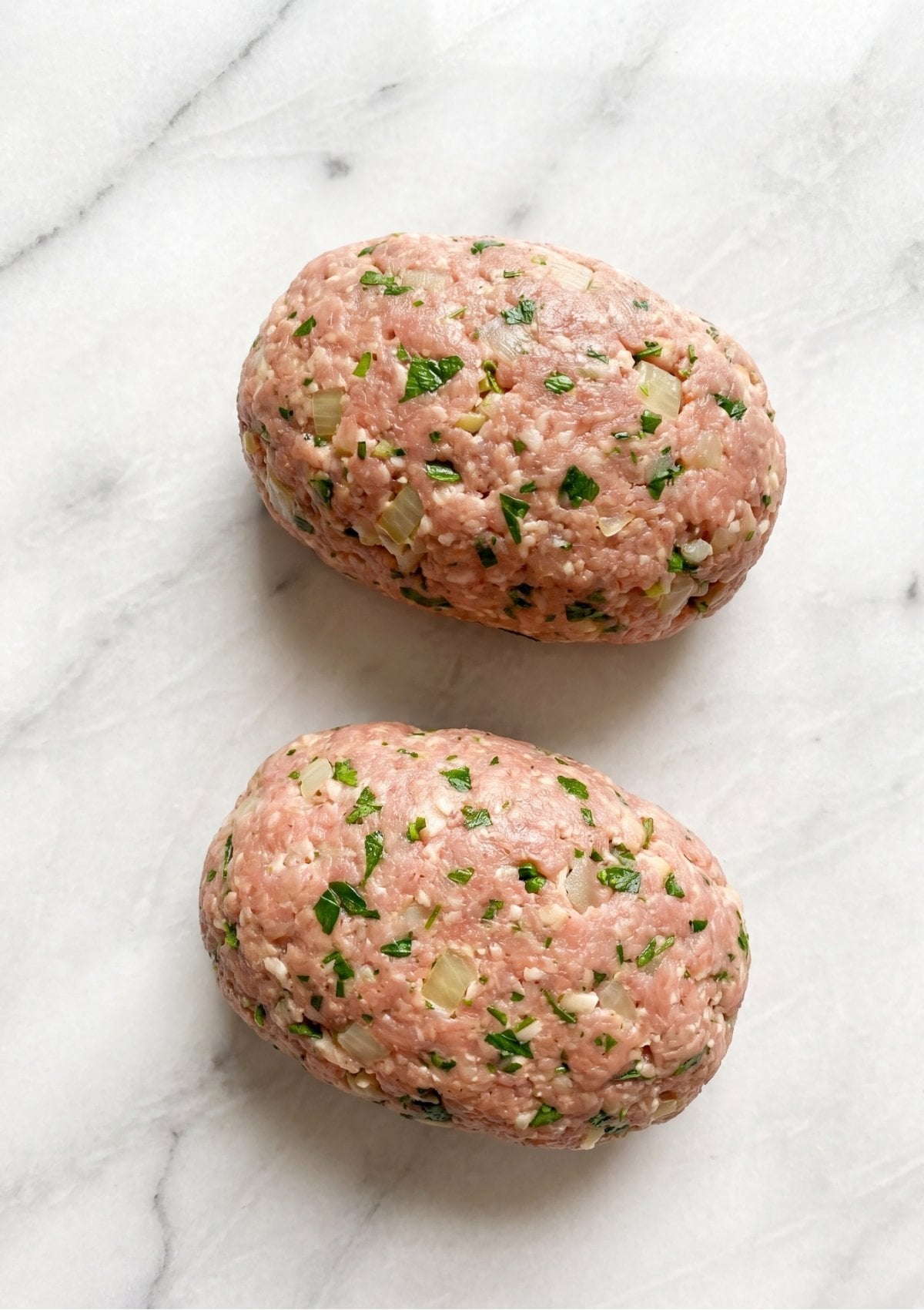 Two raw, oval-shaped ground turkey mini meatloaves mixed with visible pieces of onion and chopped parsley sitting on a white marble countertop