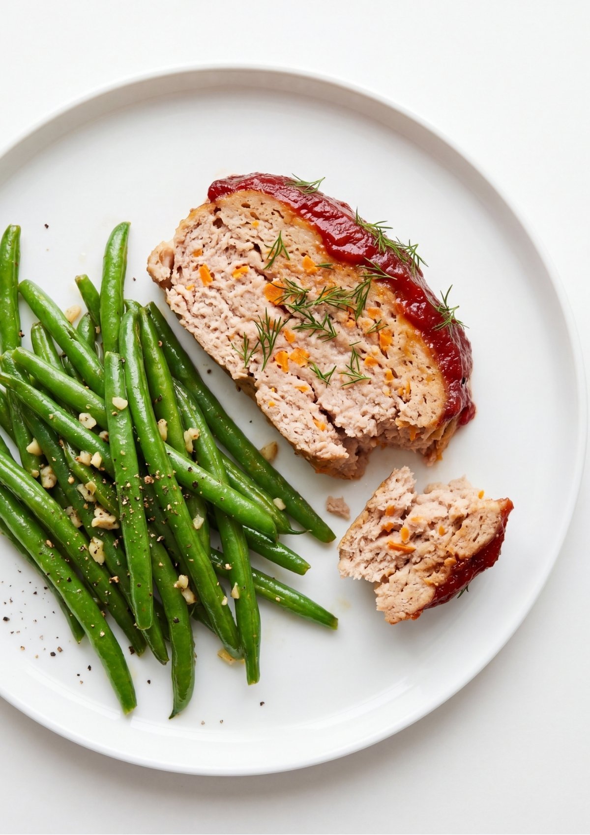 A thick slice of glazed turkey and beef meatloaf garnished with fresh dill served next to a pile of seasoned green beans on a white plate.