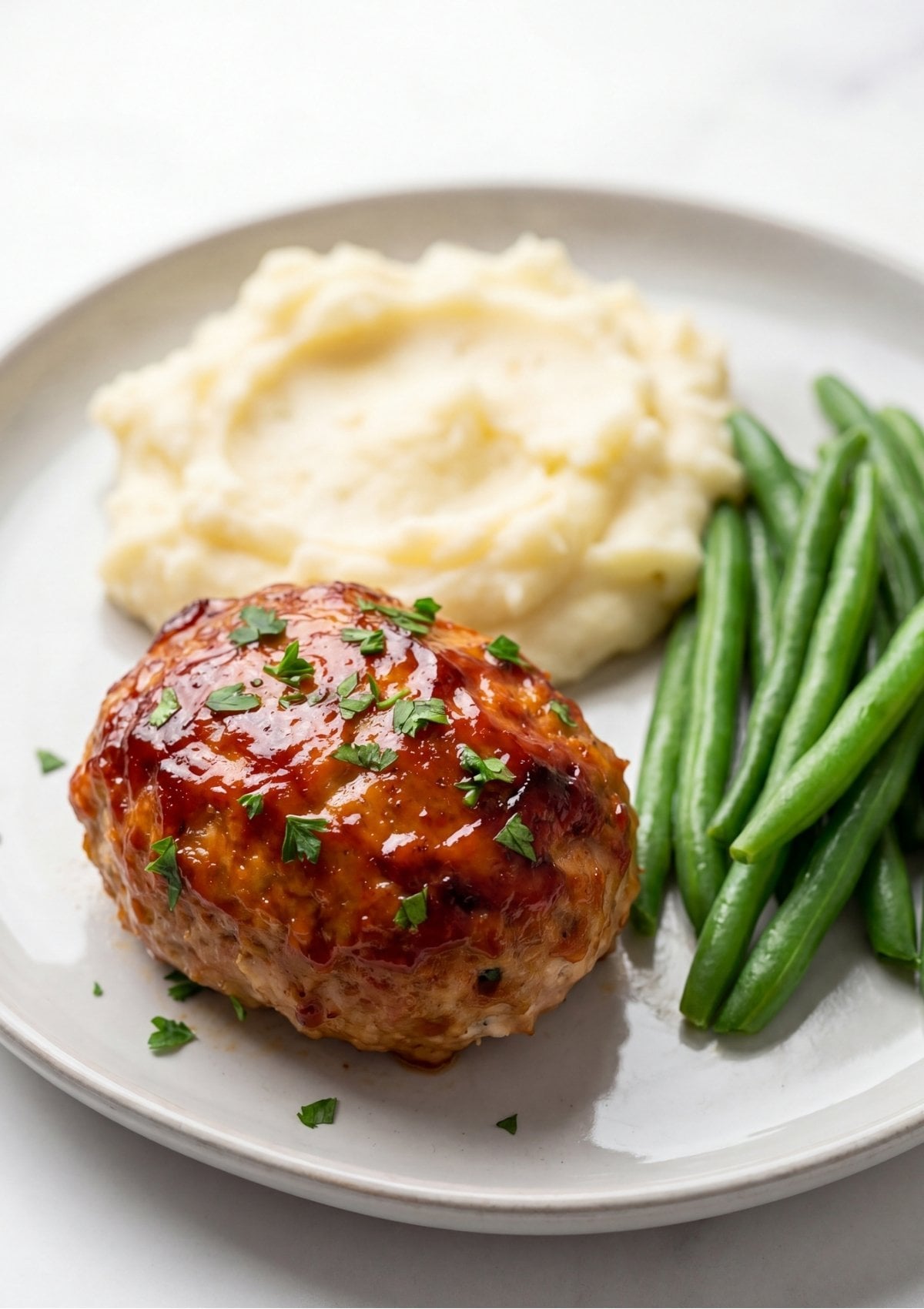 A single mini turkey meatloaf covered in a savory glaze served on a white plate alongside a scoop of mashed potatoes and green beans
