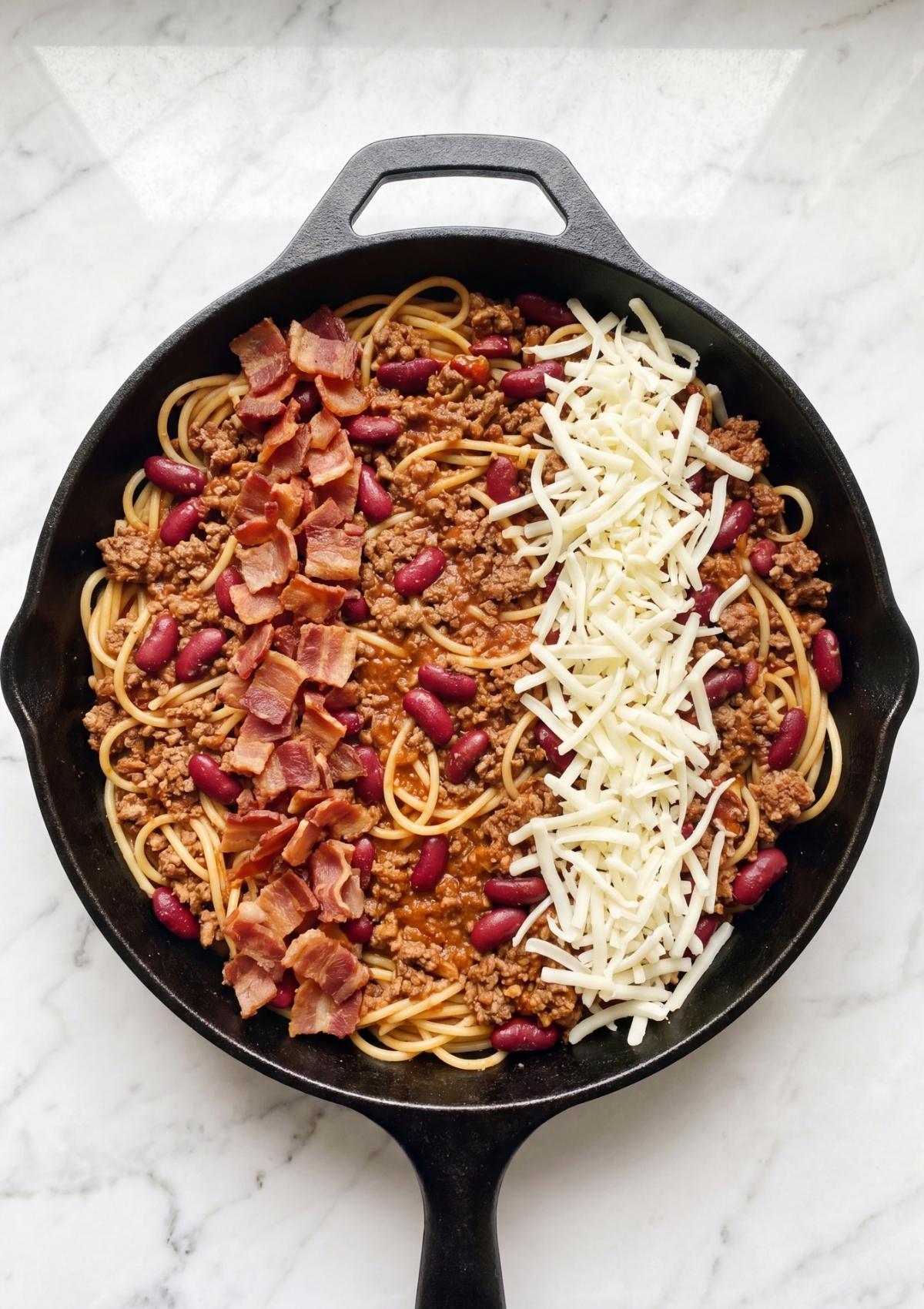 Layers of spaghetti, chili meat sauce, crispy bacon, and shredded cheese in a skillet before baking.