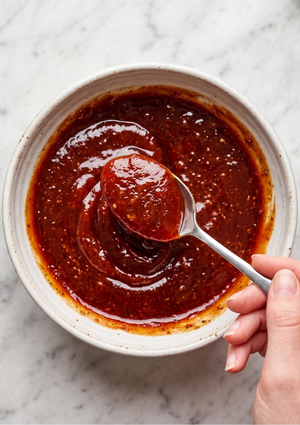 A hand using a spoon to swirl a dark red, textured Meatloaf Glaze Recipe in a small ceramic bowl.