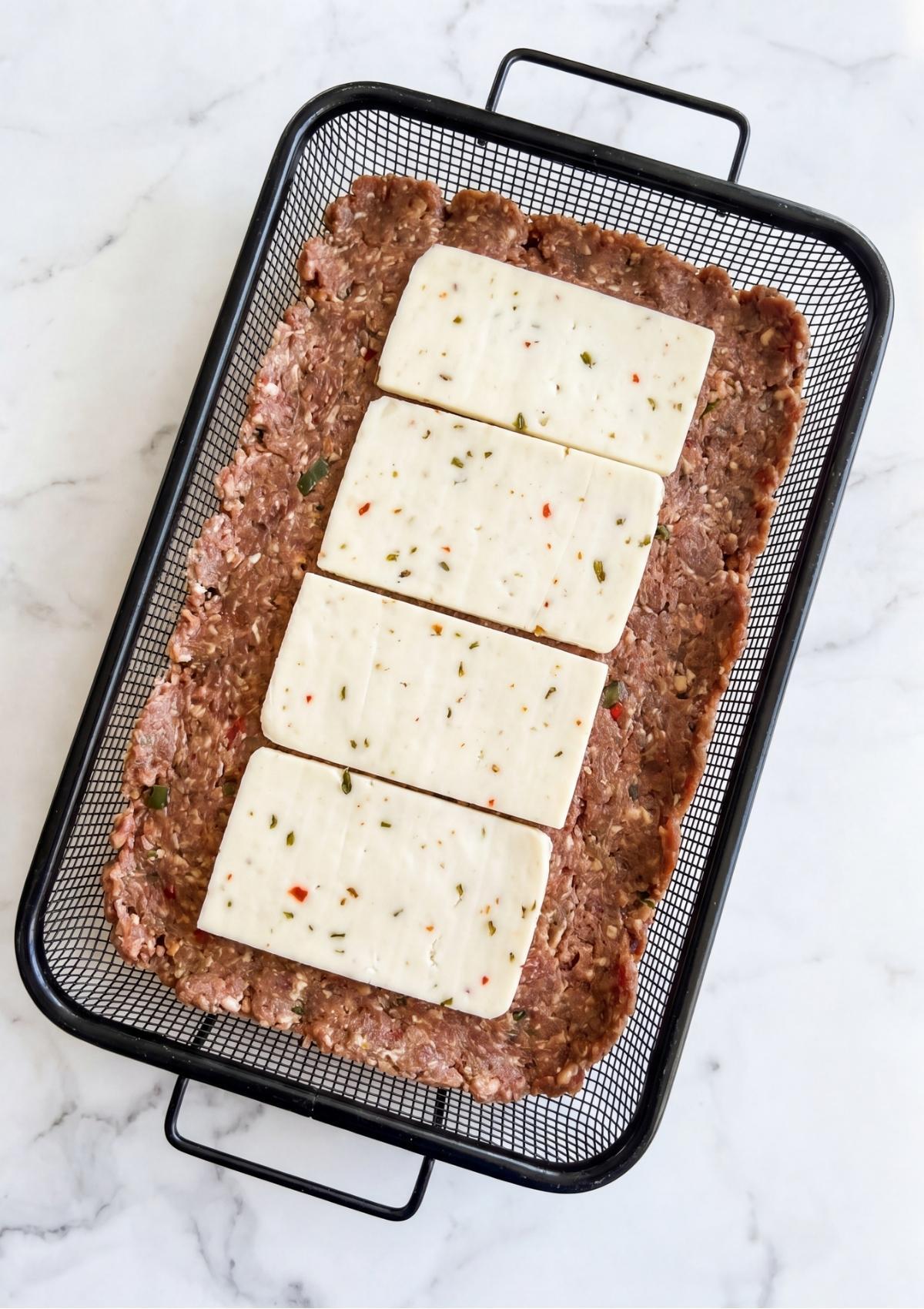 Four thick slices of pepper jack cheese layered over a flattened layer of raw meatloaf mixture in a black wire basket.