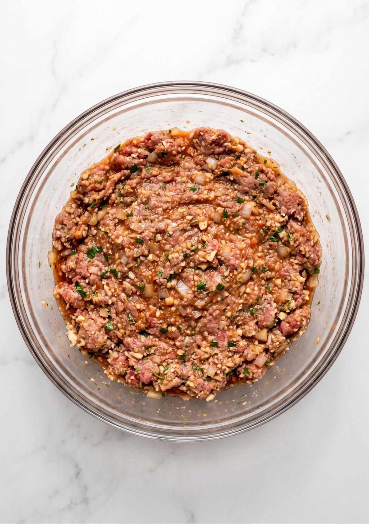 A glass bowl containing the thoroughly mixed ground meat, herbs, and aromatics, ready to be shaped.
