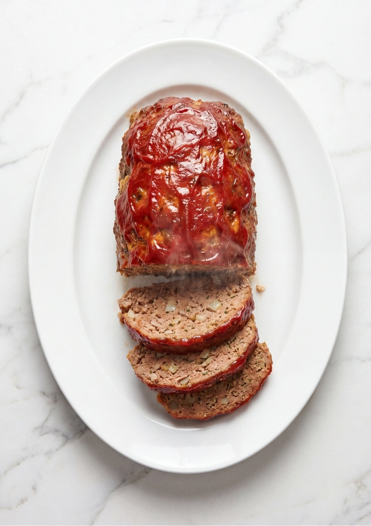 A whole meatloaf topped with a thick red tomato glaze on a white oval platter, with three slices cut and layered at the bottom showing a moist texture and diced onions inside.