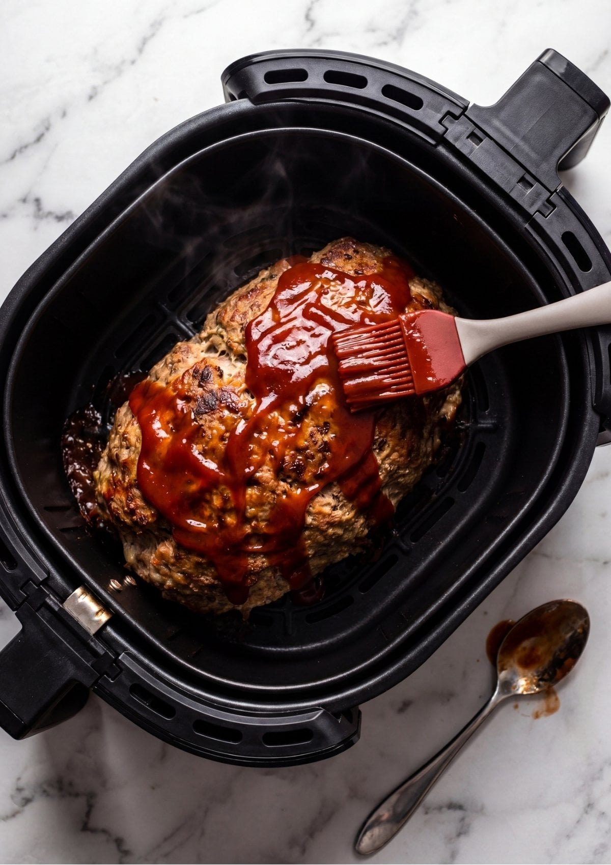 A hand using a red silicone brush to spread a thick red glaze over a meatloaf inside an air fryer.
