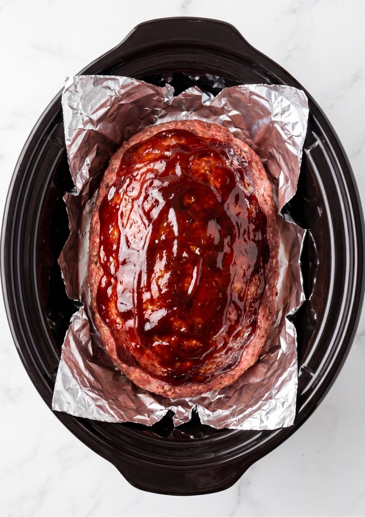 An unbaked meatloaf in a slow cooker with a layer of red glaze spread across the top