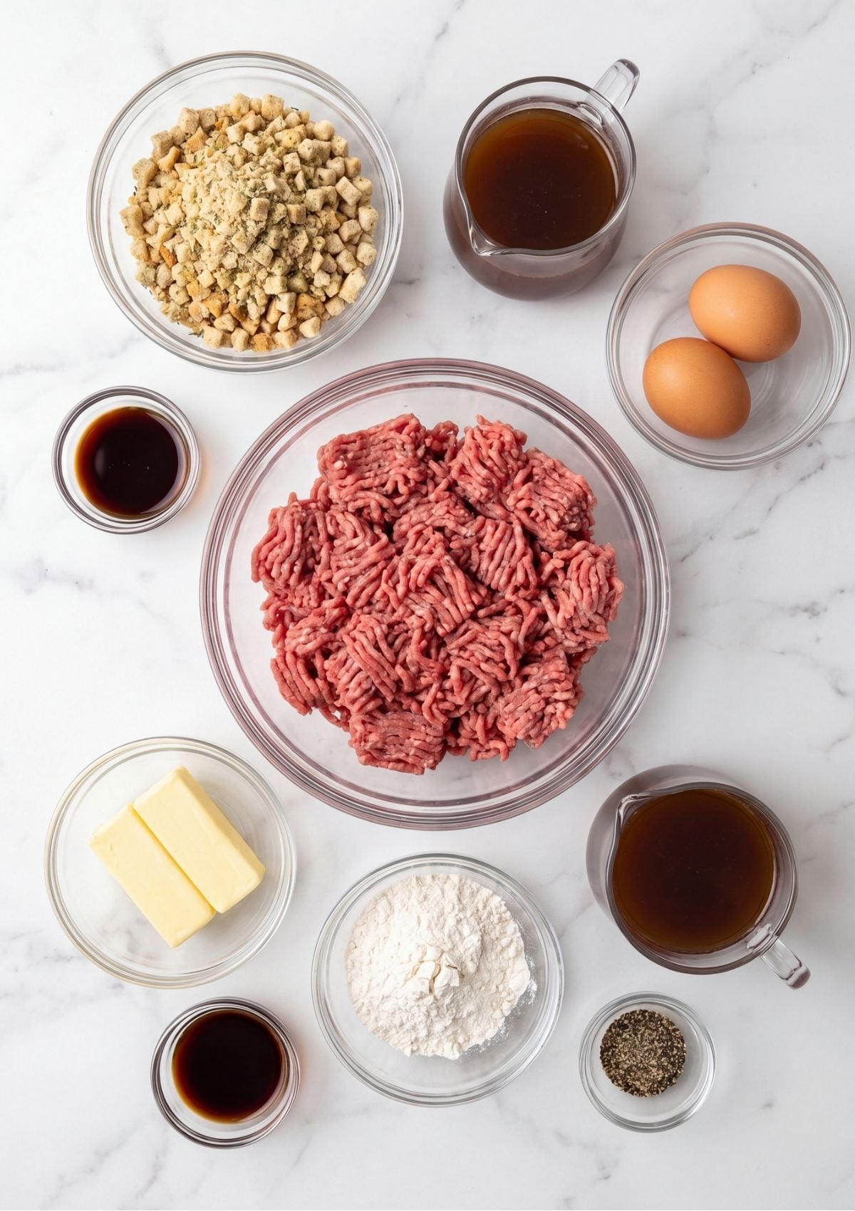 An overhead view of ingredients including ground beef, eggs, butter, flour, broth, and stuffing mix in small bowls