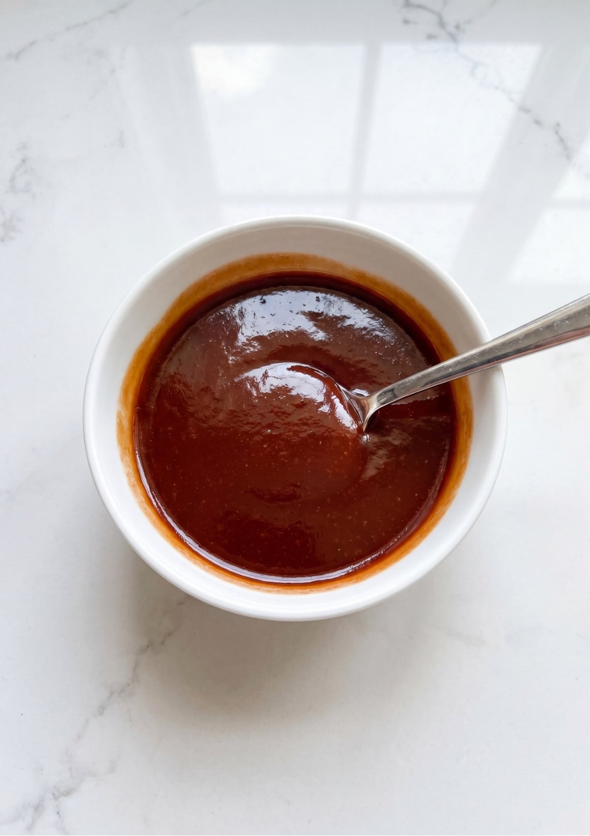 A small white bowl filled with a smooth reddish brown meatloaf glaze and a silver spoon