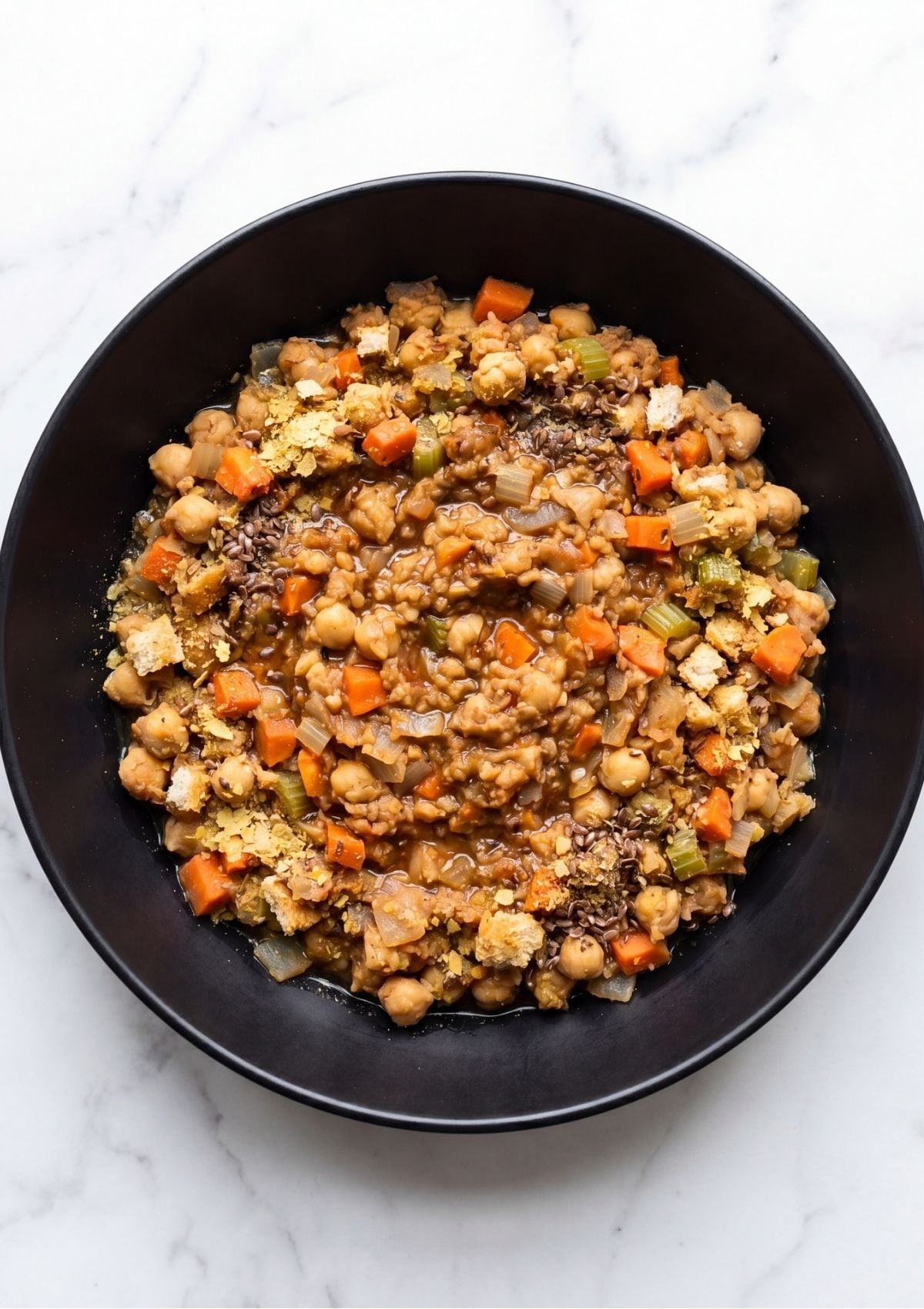 A black bowl containing a mixture of mashed chickpeas, diced carrots, celery, and seasonings for a vegan meatloaf