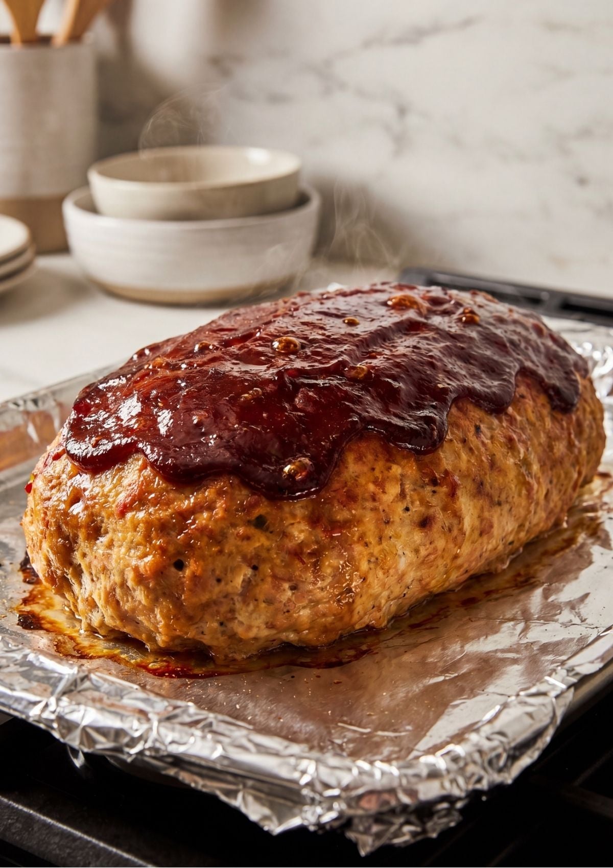 A fully cooked, steaming turkey meatloaf topped with a dark red glaze on a foil-lined baking sheet.