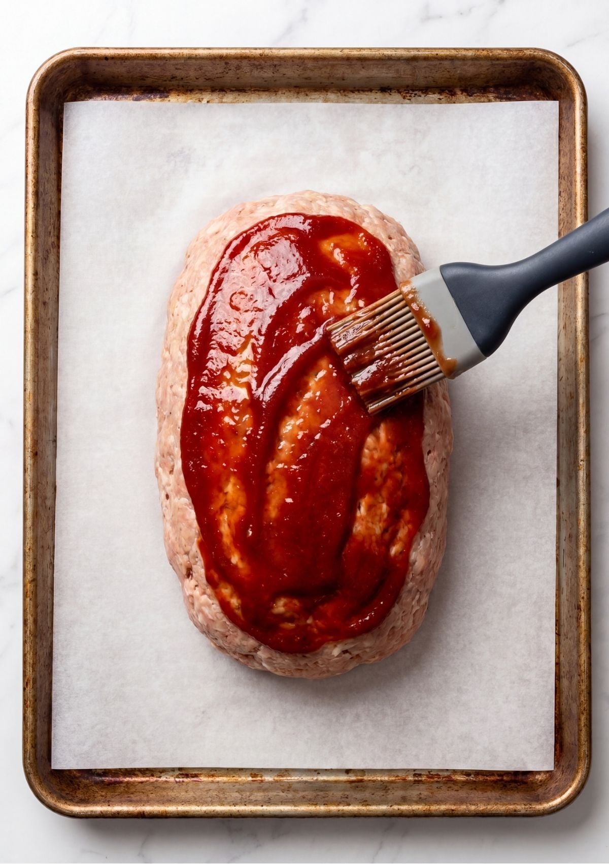 A grey silicone brush spreading red ketchup glaze over the top of a raw ground turkey meatloaf on a parchment lined baking sheet.