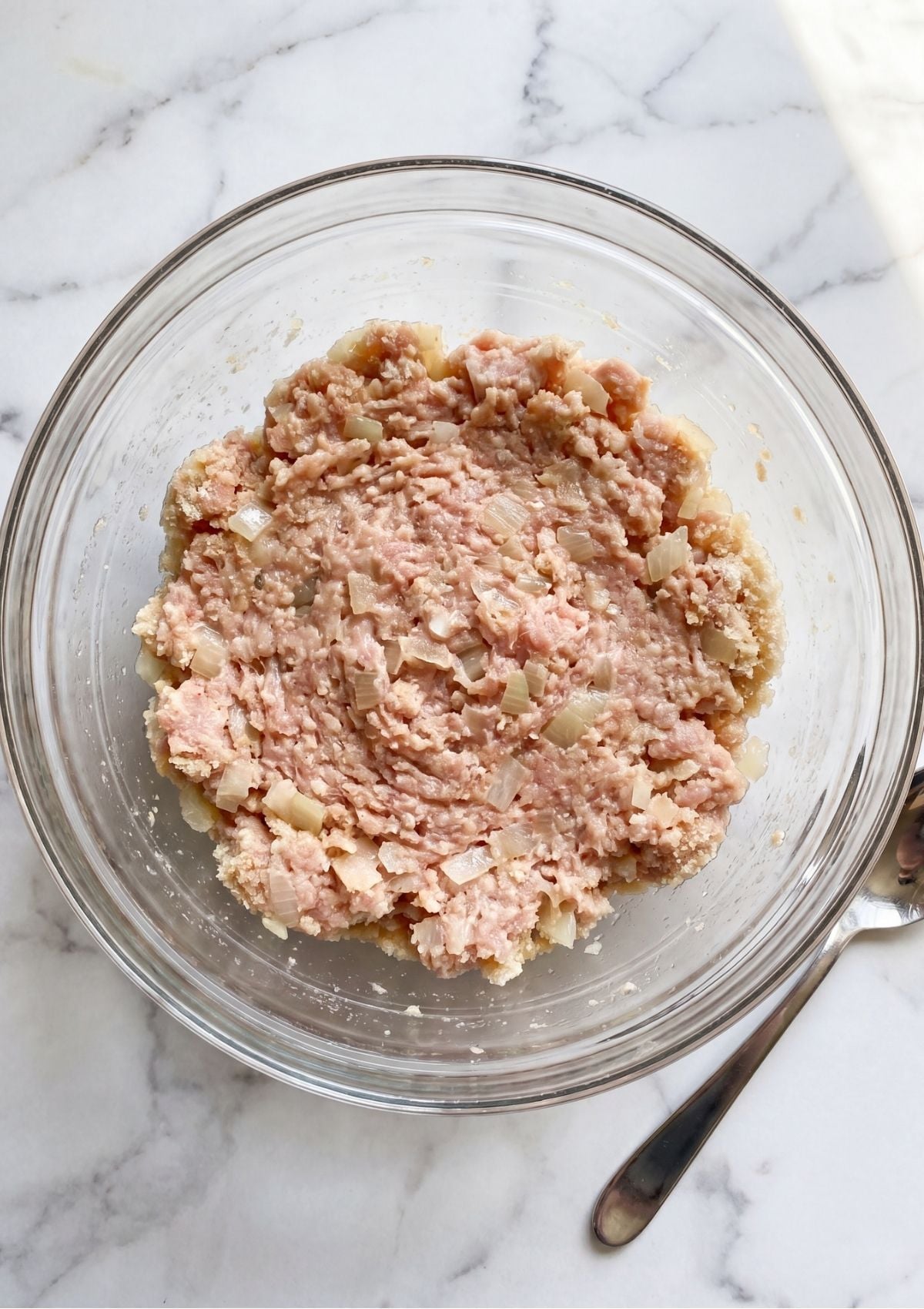 Raw ground turkey mixed with diced onions and breadcrumbs in a clear glass bowl on a marble countertop.