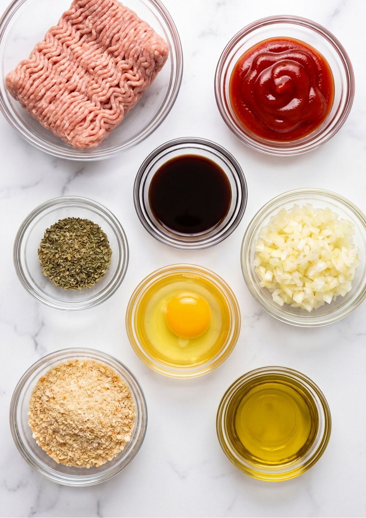 Overhead view of measured ingredients for turkey meatloaf including ground turkey, ketchup, Worcestershire sauce, chopped onions, an egg, breadcrumbs, and olive oil in small glass bowls