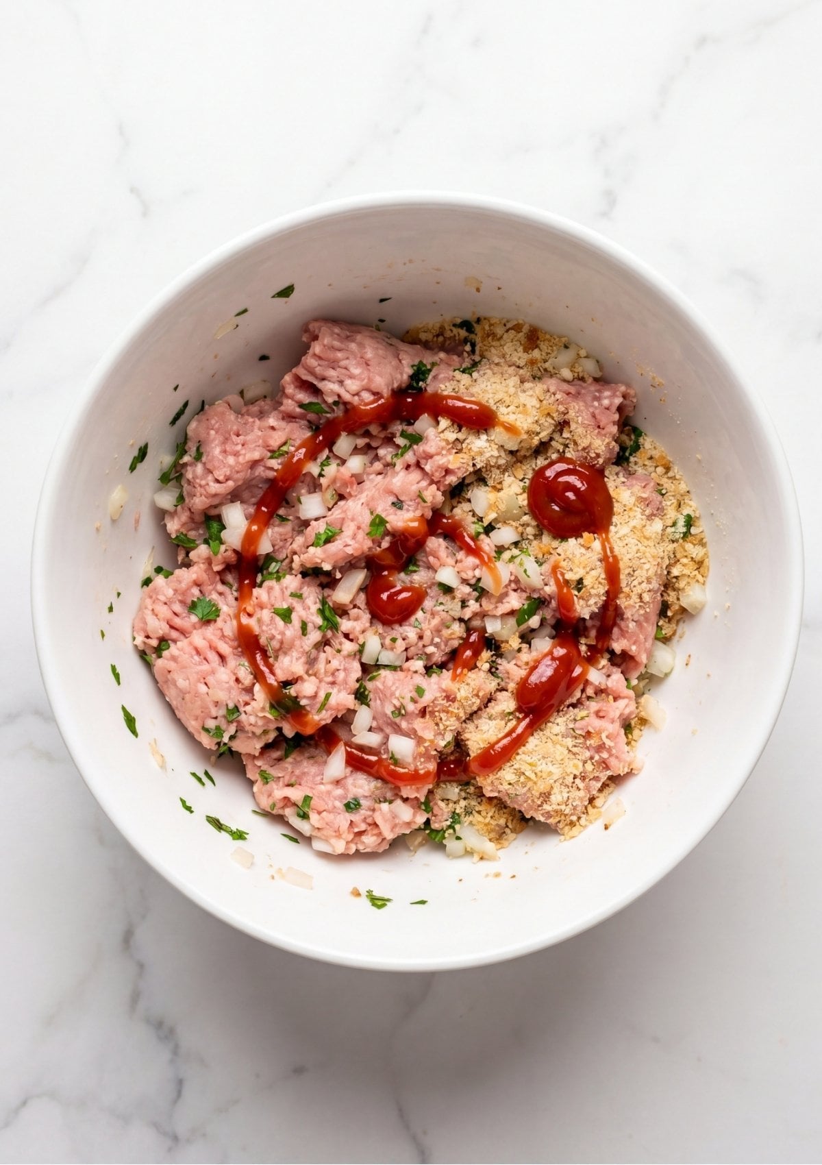 Ground turkey being mixed with breadcrumbs, diced onions, chopped fresh parsley, and a drizzle of ketchup inside a white ceramic mixing bowl