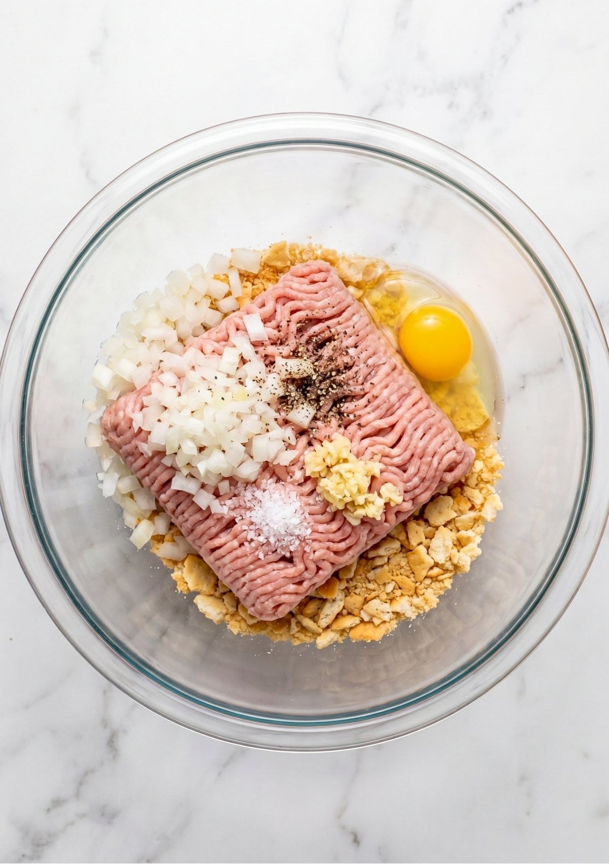 Unmixed ingredients including ground turkey, diced onions, a whole egg, minced garlic, and crushed crackers in a glass mixing bowl