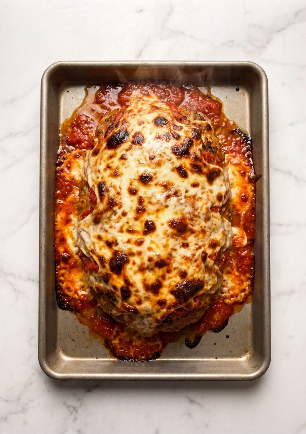 A whole baked turkey meatloaf fresh out of the oven on a metal baking sheet, completely covered in melted, bubbly, golden brown mozzarella cheese and red marinara sauce
