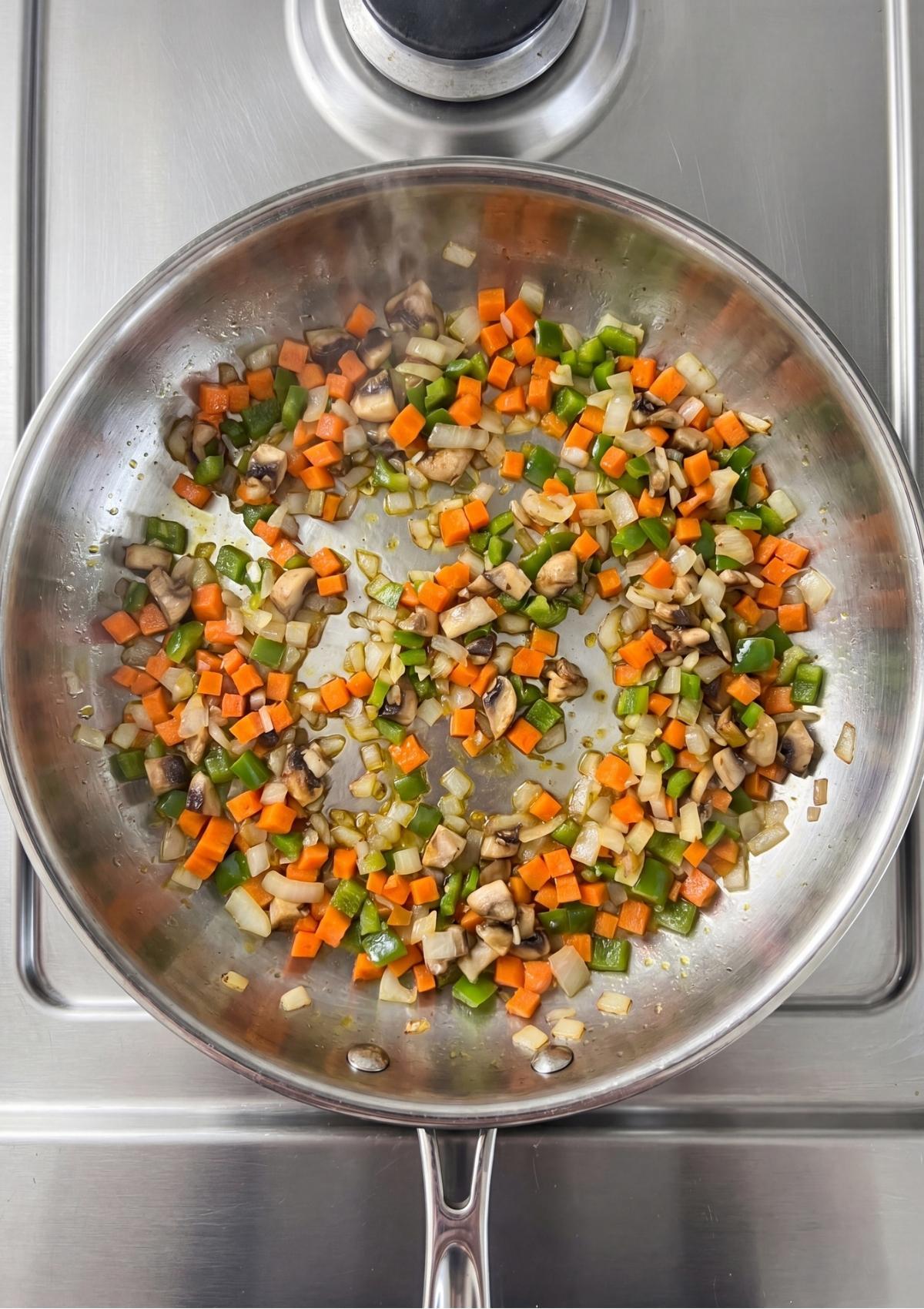 Finely diced carrots, onions, green bell peppers, and mushrooms being sautéed in a stainless steel skillet on the stove.