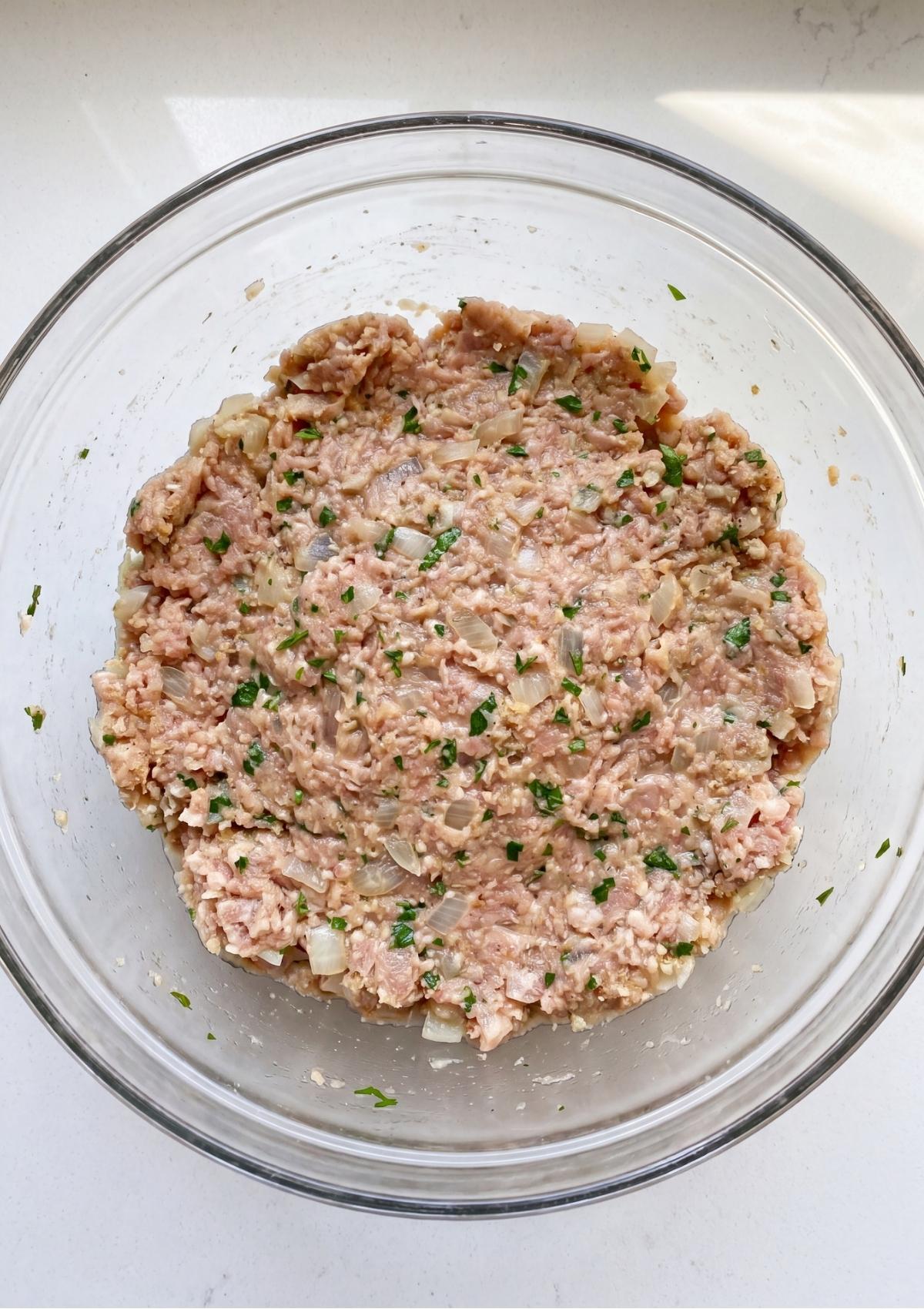 A large glass bowl filled with the ground turkey meatloaf mixture combined with herbs, breadcrumbs, and sautéed onions