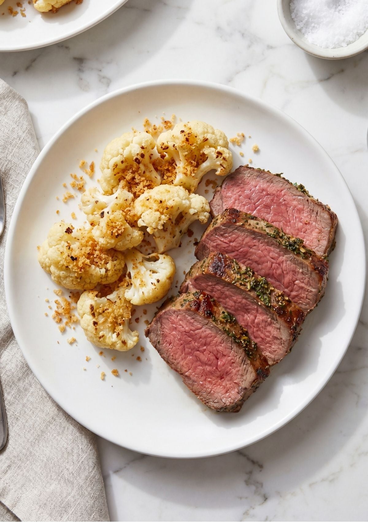 Overhead view of a white dinner plate containing four thick slices of medium-rare roast beef and a side of golden breaded roasted cauliflower florets