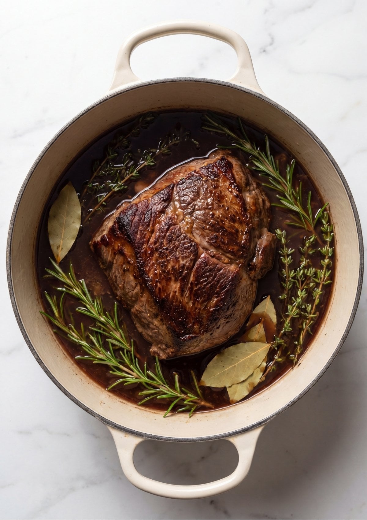 Seared beef roast resting in a dark red wine and broth braising liquid inside a Dutch oven with fresh rosemary sprigs, thyme, and bay leaves before cooking.