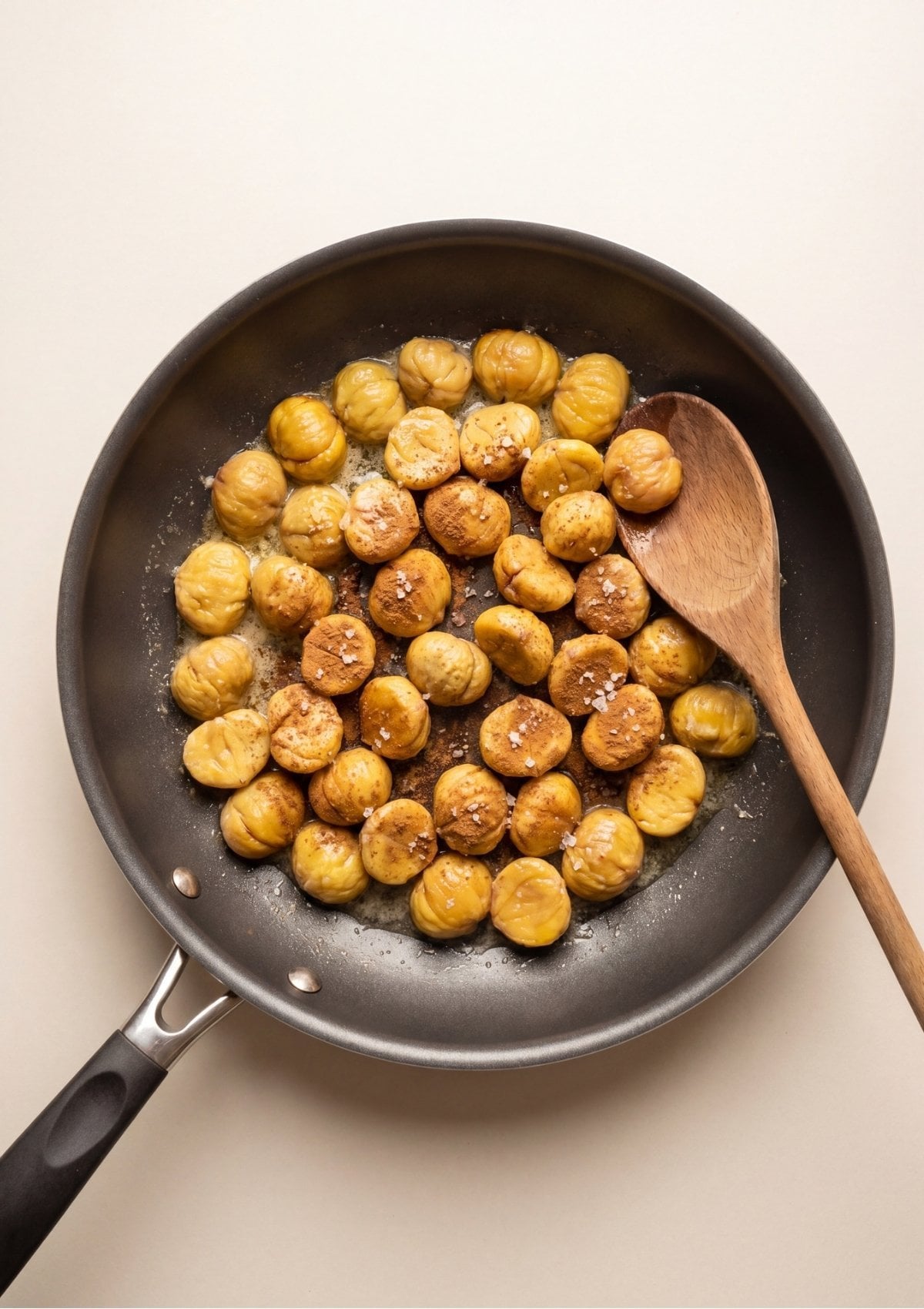 Golden peeled chestnuts being tossed with melted butter, ground cinnamon, and sea salt in a black skillet with a wooden spoon.