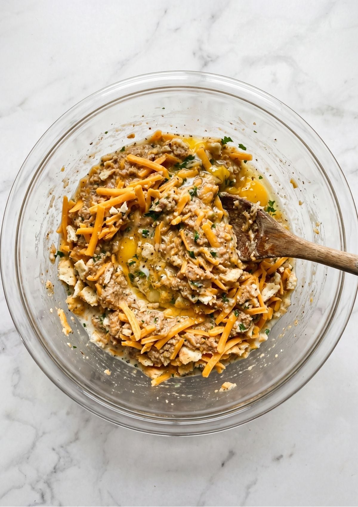 A glass bowl containing crushed crackers, shredded cheese, and egg being combined for a meatloaf recipe.