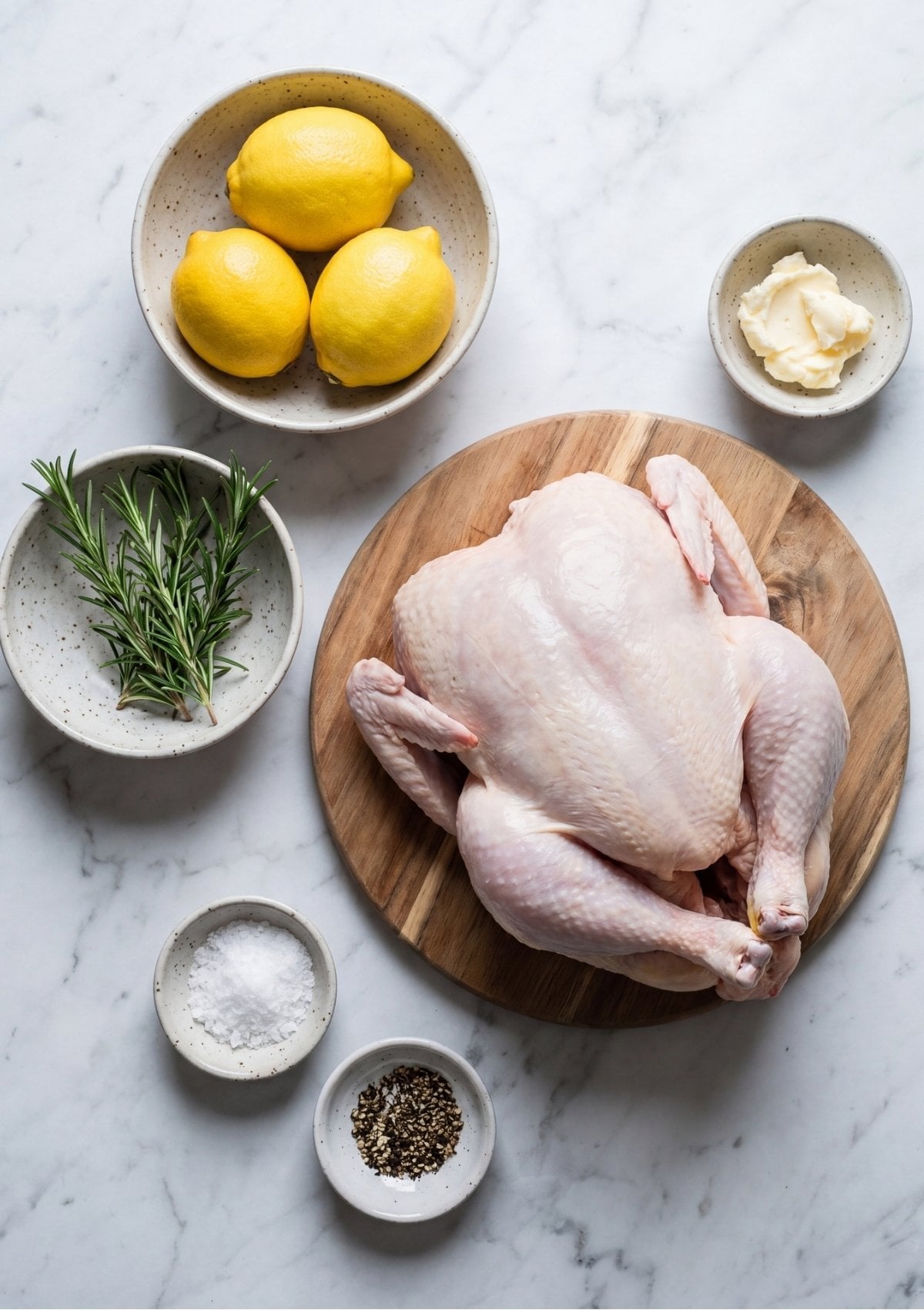 Whole raw chicken on a wooden cutting board surrounded by small bowls containing lemons, softened butter, fresh rosemary, sea salt, and black pepper