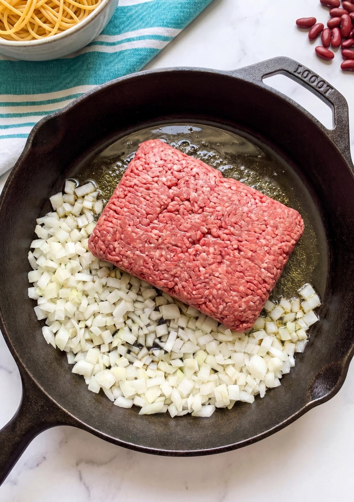 Raw ground beef and diced white onions cooking in a cast iron skillet on a marble surface.