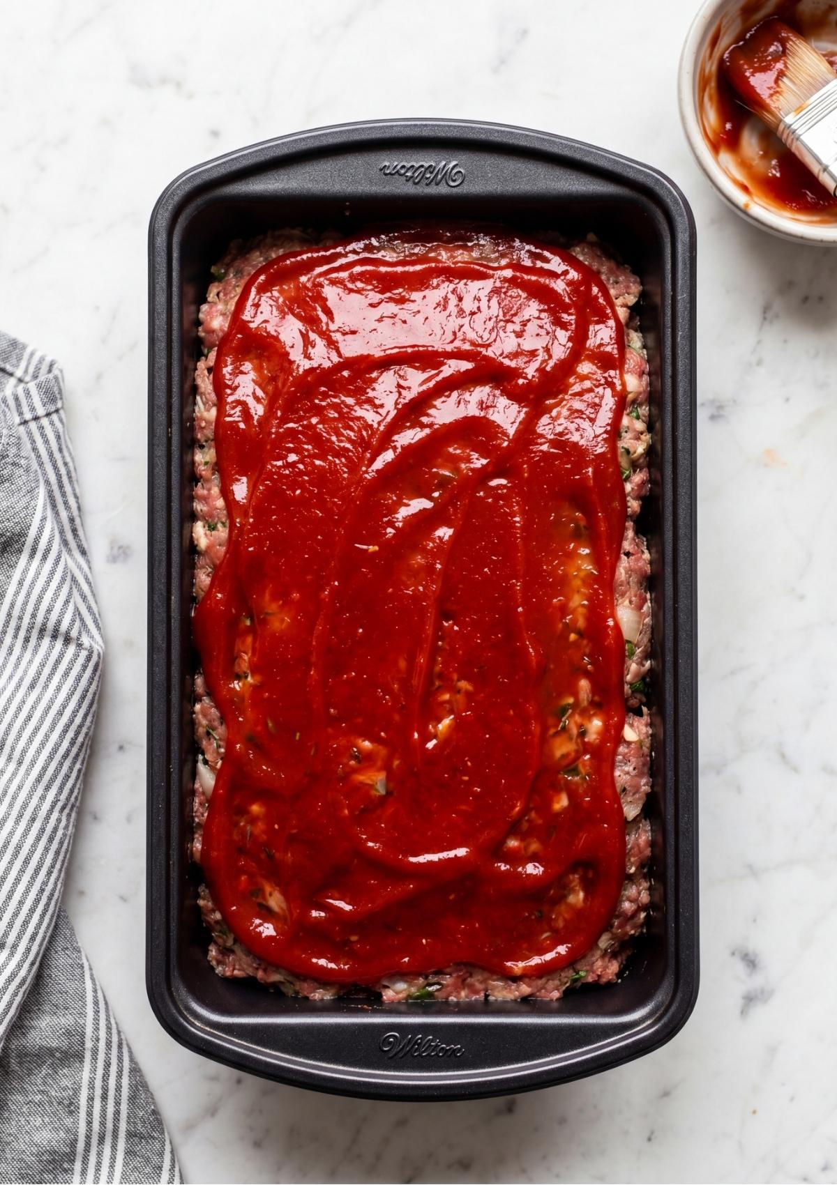 Uncooked meatloaf in a pan topped with a generous, smooth layer of red glaze.