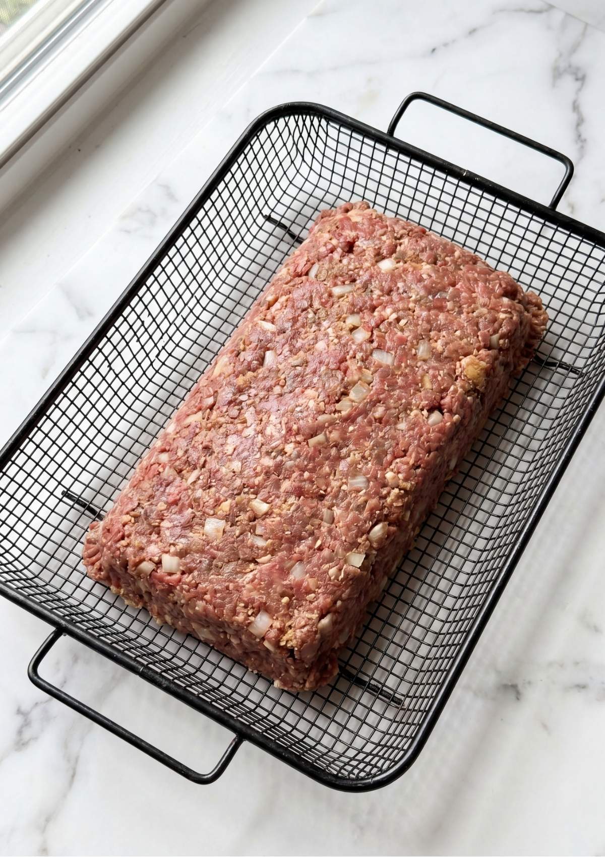 A perfectly shaped raw meatloaf loaf sitting in a black wire mesh basket, showing visible bits of onion and seasoning.
