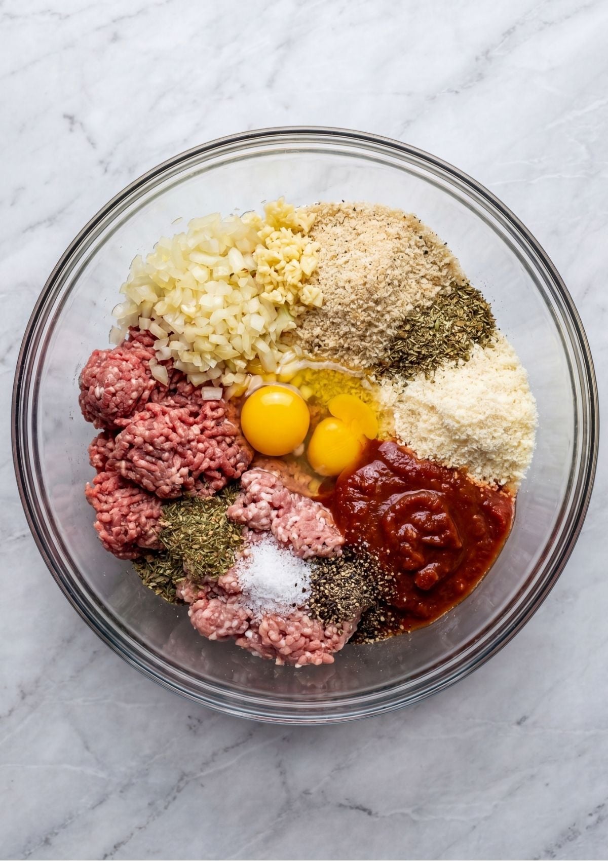 A glass mixing bowl filled with raw ground meat, eggs, breadcrumbs, minced garlic, onions, herbs, and marinara sauce.