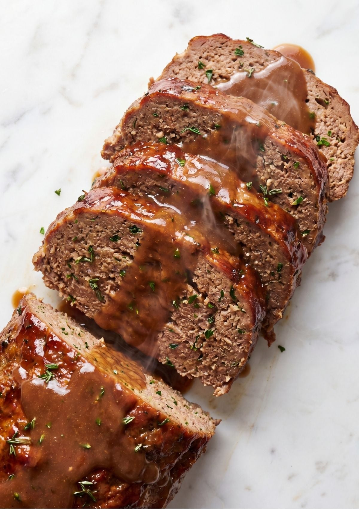 Close up of thick slices of meatloaf drizzled with glistening brown gravy on a white marble surface