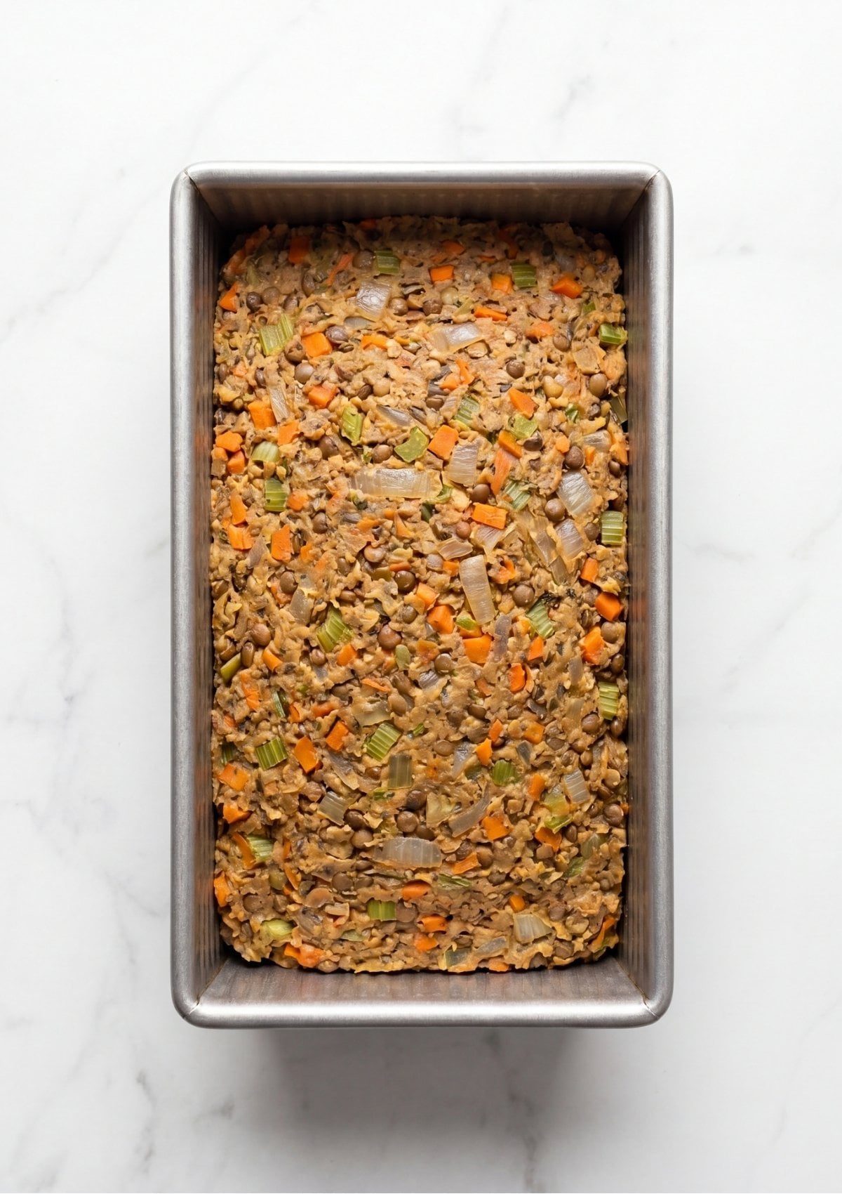 A top down view of raw vegan meatloaf mixture with visible chickpeas and vegetables pressed firmly into a metal loaf pan
