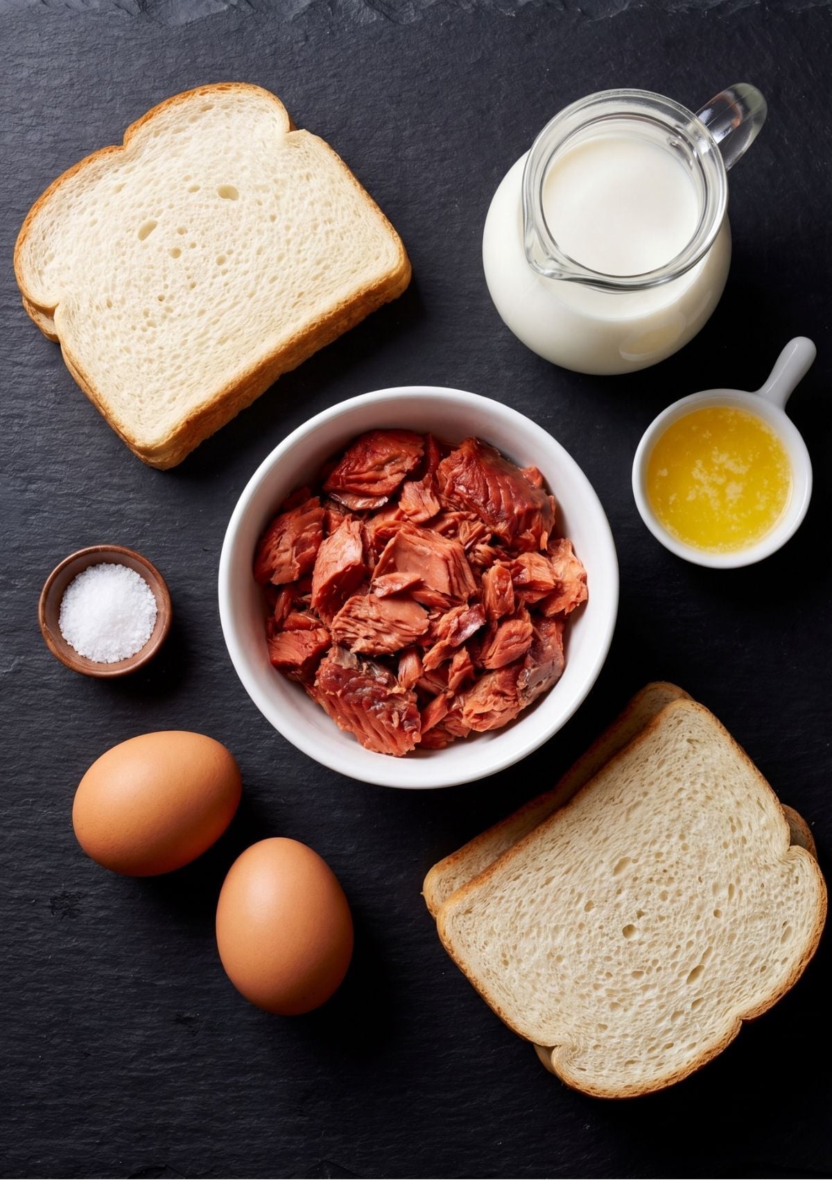 Top down view of salmon meatloaf ingredients including canned red salmon in a white bowl, slices of white bread, two brown eggs, milk in a glass jug, melted butter, and a small bowl of salt on a dark slate background