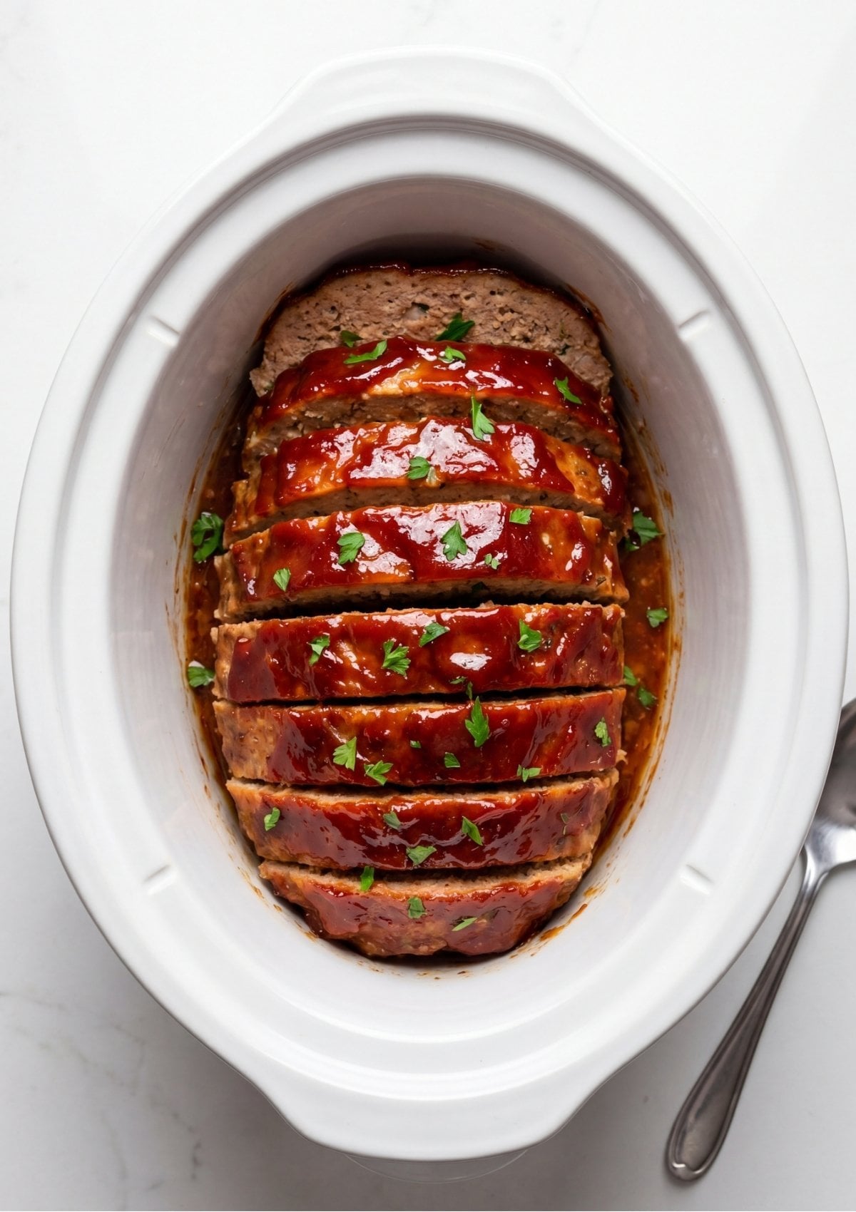 A sliced turkey meatloaf glazed with a glossy red sauce and garnished with fresh green parsley inside a white slow cooker insert beside a serving spoon