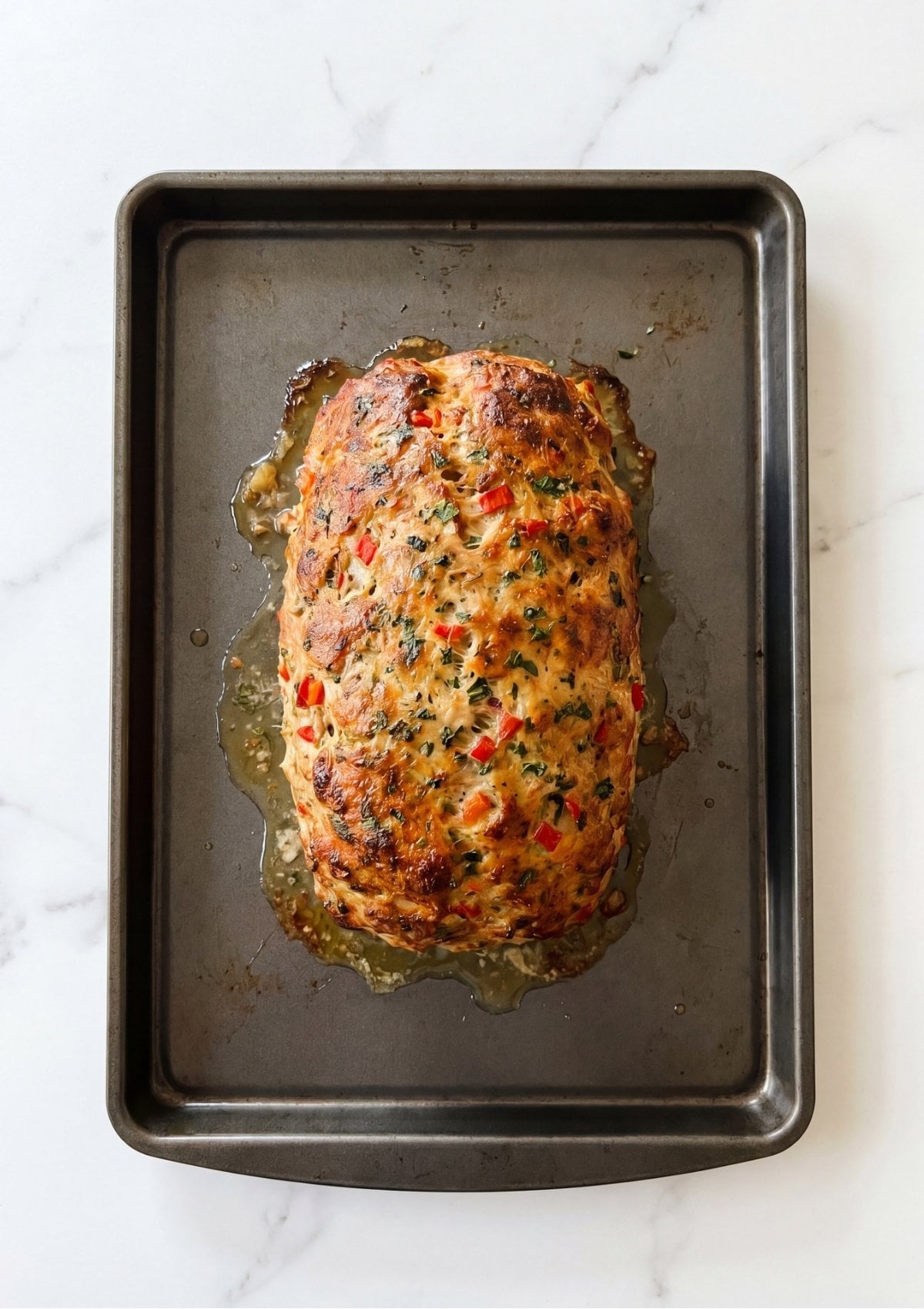 A top down view of a whole baked turkey meatloaf showing visible flecks of red bell pepper and green parsley on a dark metal baking sheet
