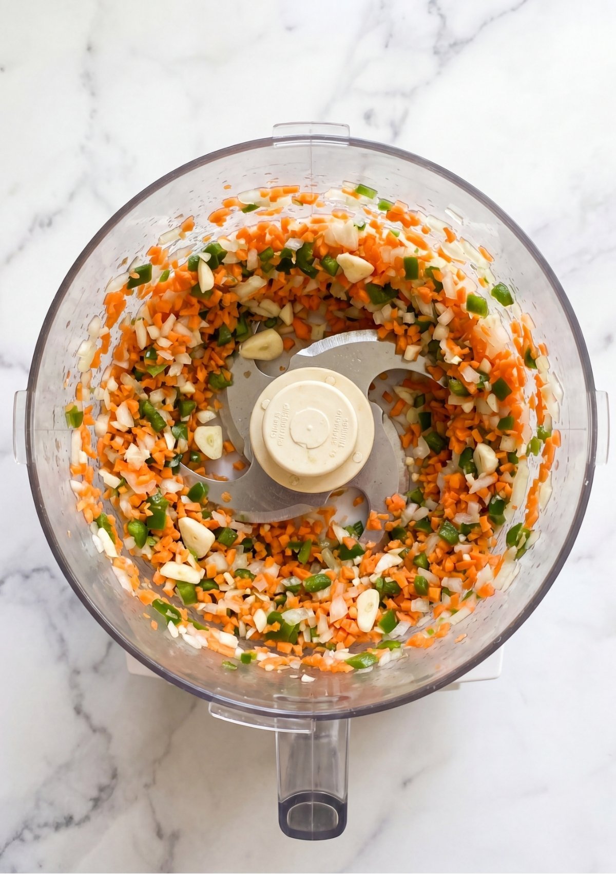 Finely chopped carrots, onions, garlic, and green bell peppers inside the clear bowl of a food processor.