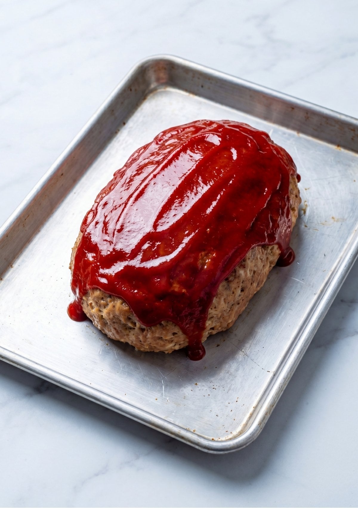 The partially baked turkey meatloaf on a sheet pan with thick rows of red glaze spread across the top.