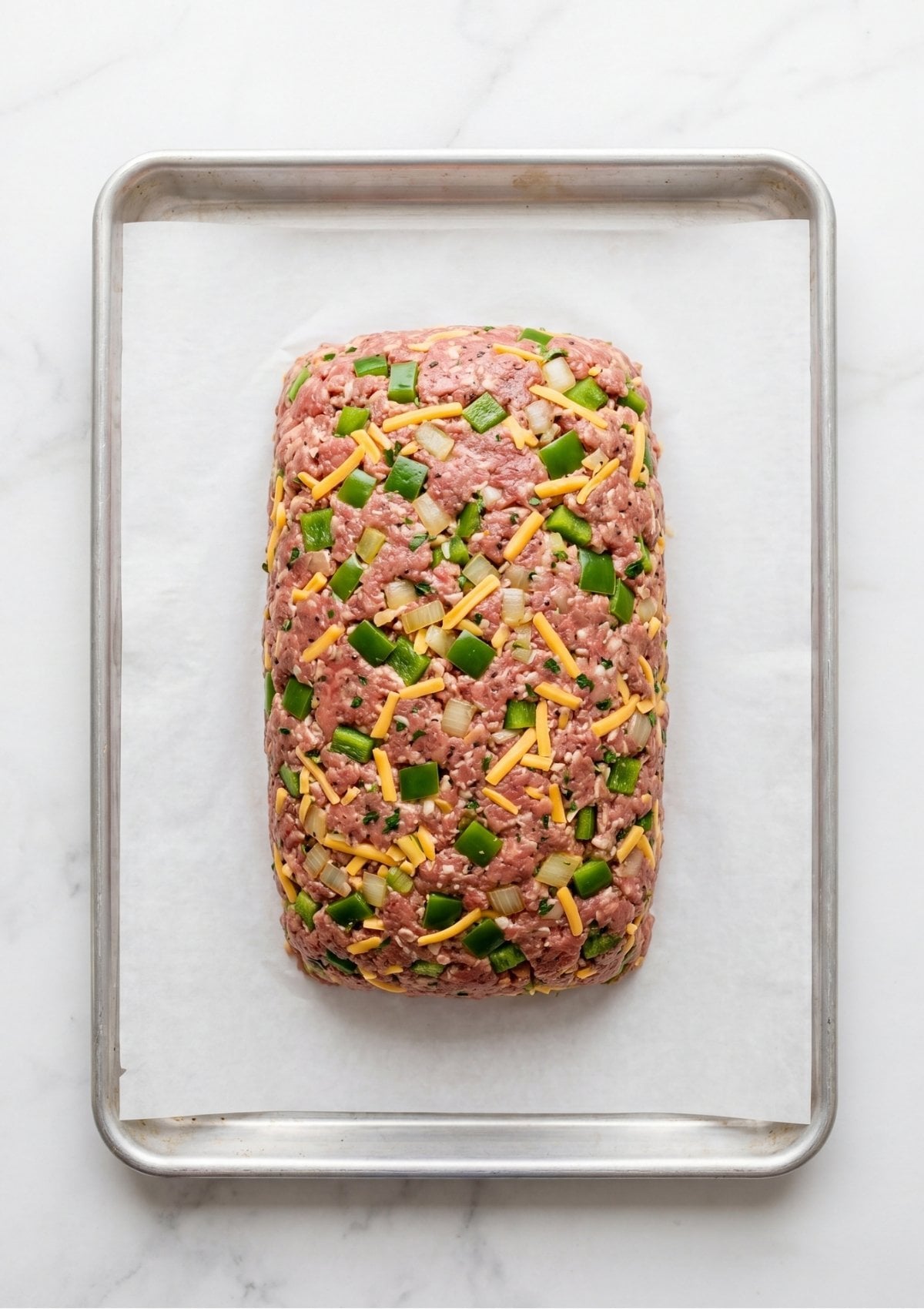 A raw mixture of beef, peppers, and cheddar cheese shaped into a rectangular loaf on a parchment-lined pan.
