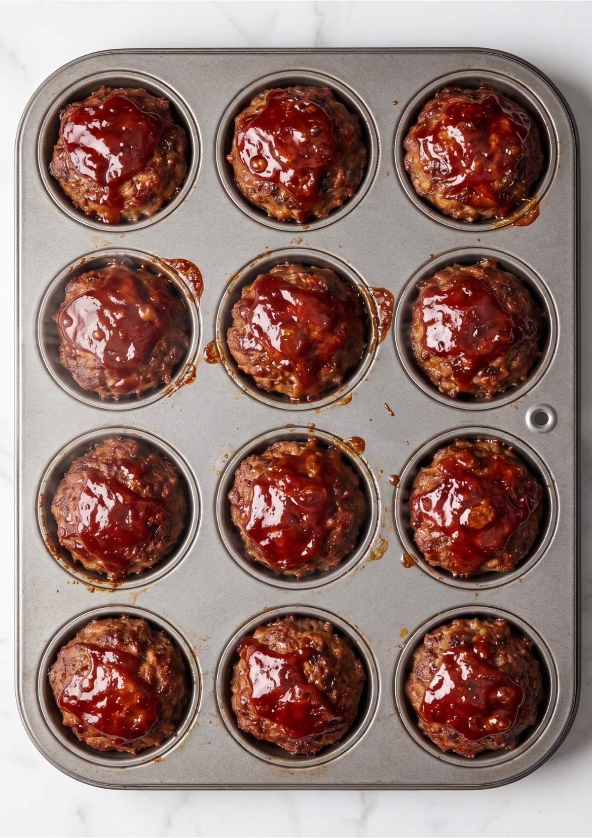 Twelve mini meatloaves topped with red glaze in a dark metal muffin tin.