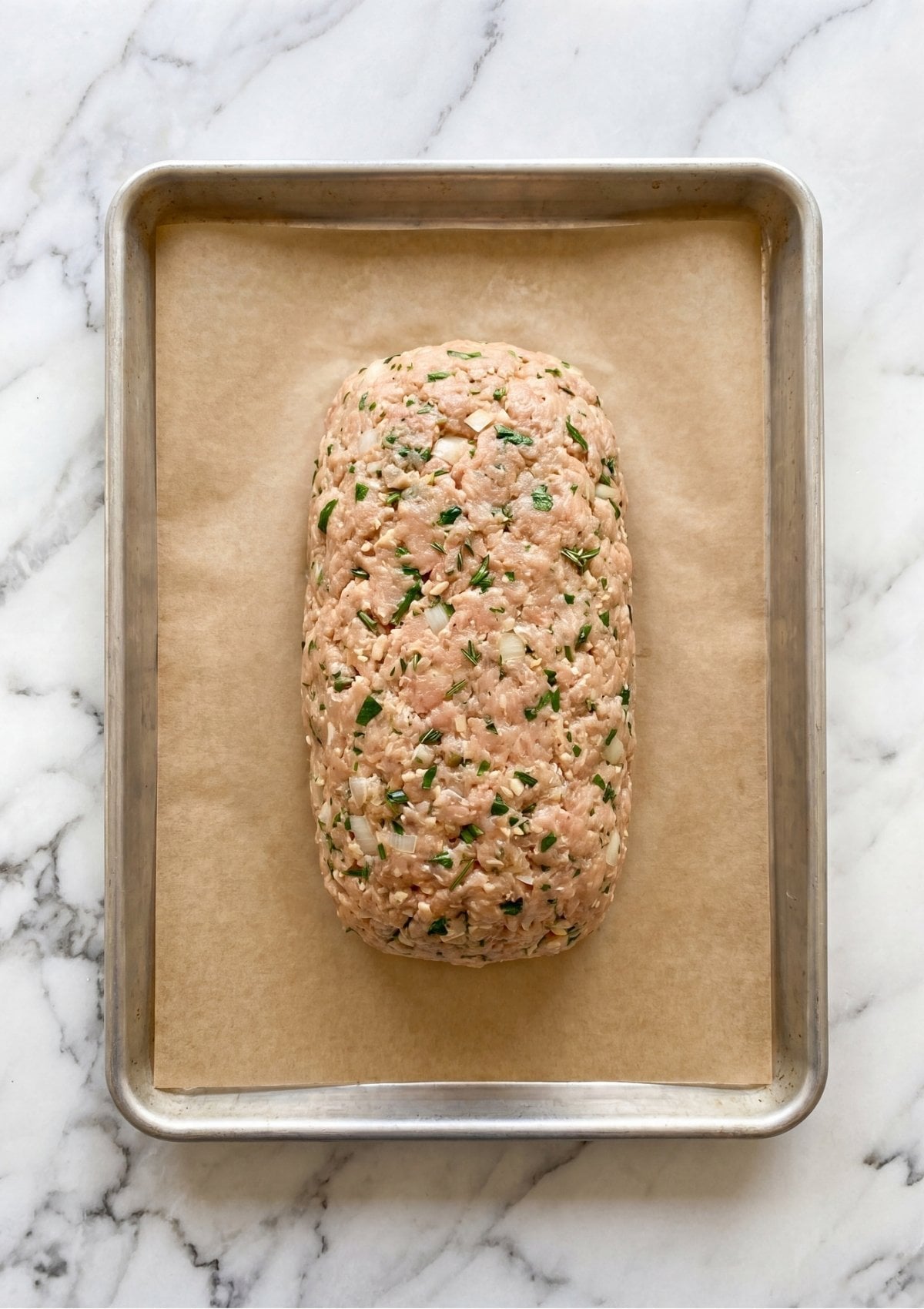 A raw, seasoned ground chicken loaf mixed with fresh herbs and onions on a parchment-lined baking sheet.