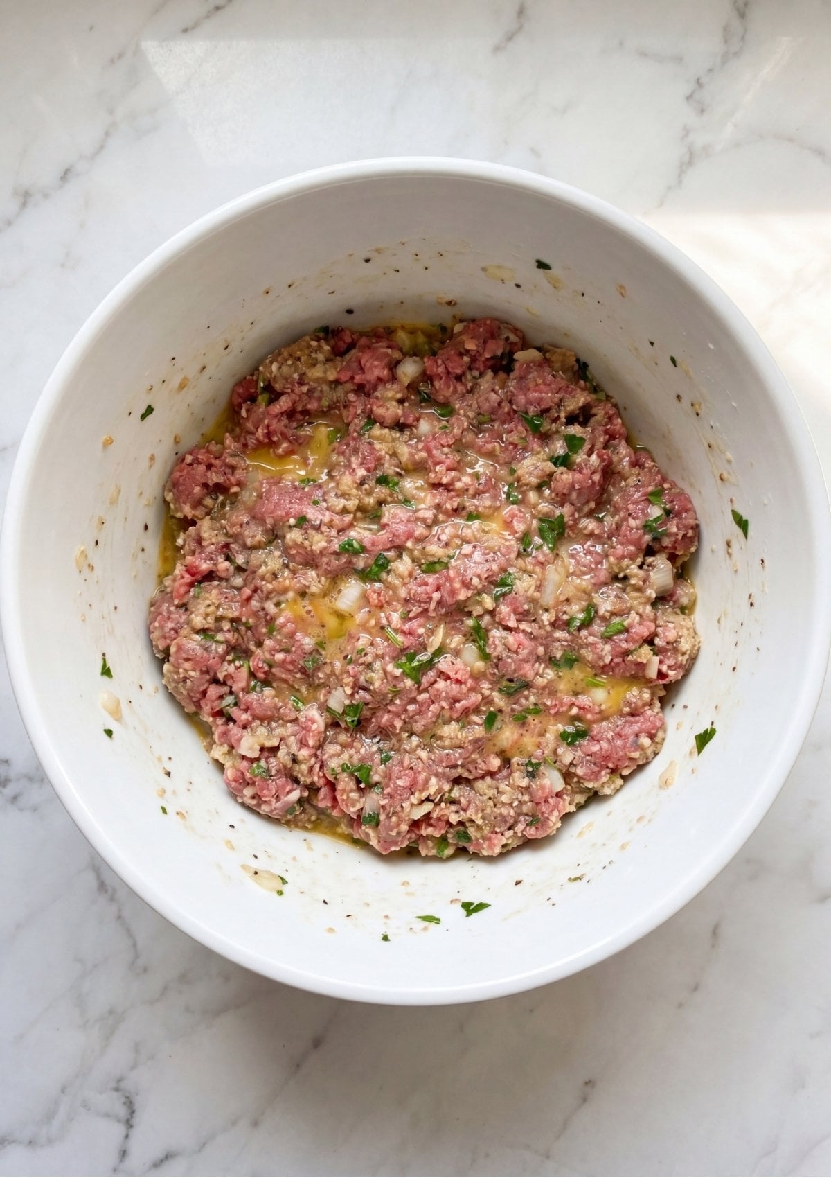 A large white mixing bowl filled with raw ground beef mixed with eggs, herbs, and breadcrumbs.