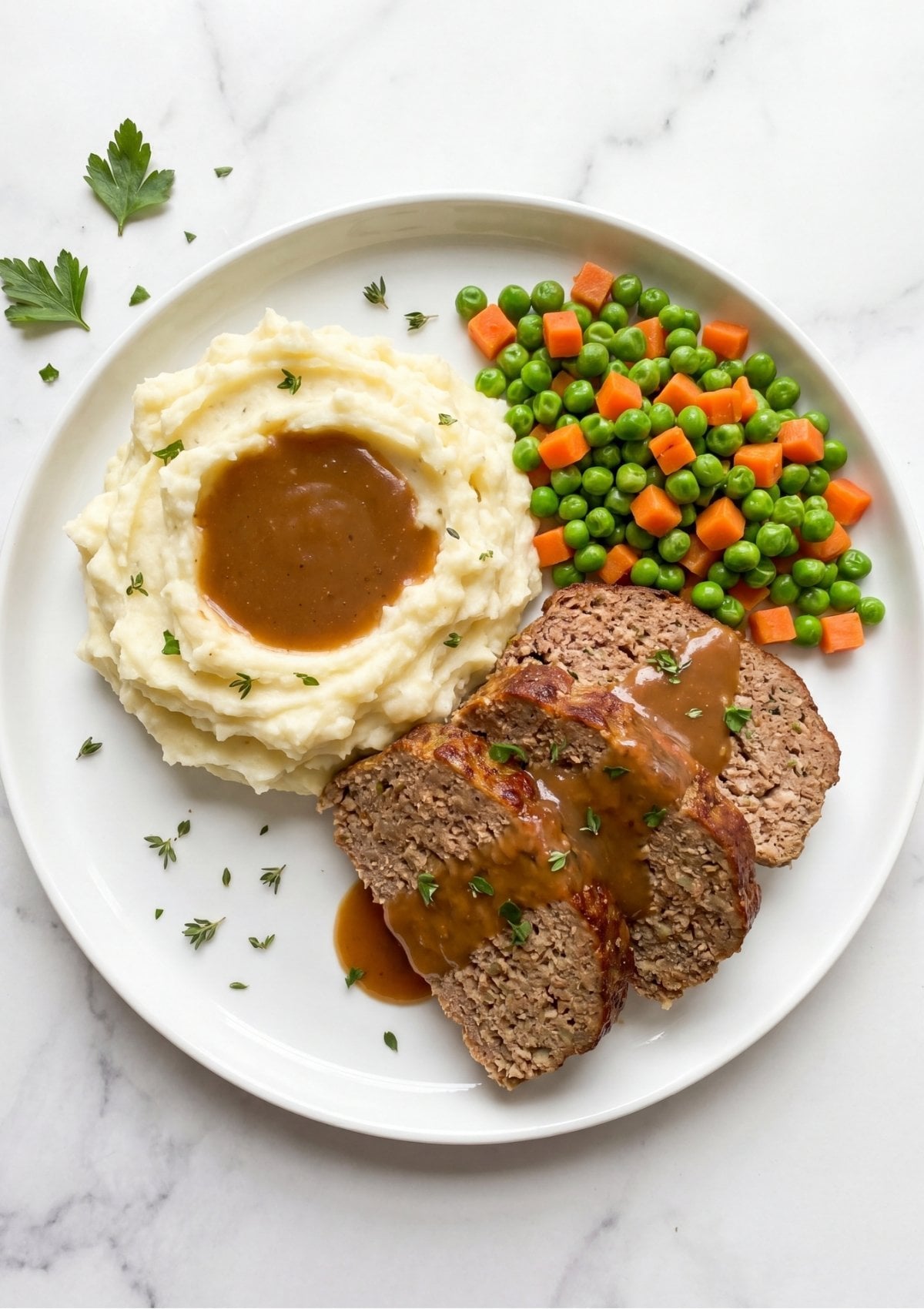A white plate featuring three slices of meatloaf with gravy, a mound of mashed potatoes, and a side of peas and carrots