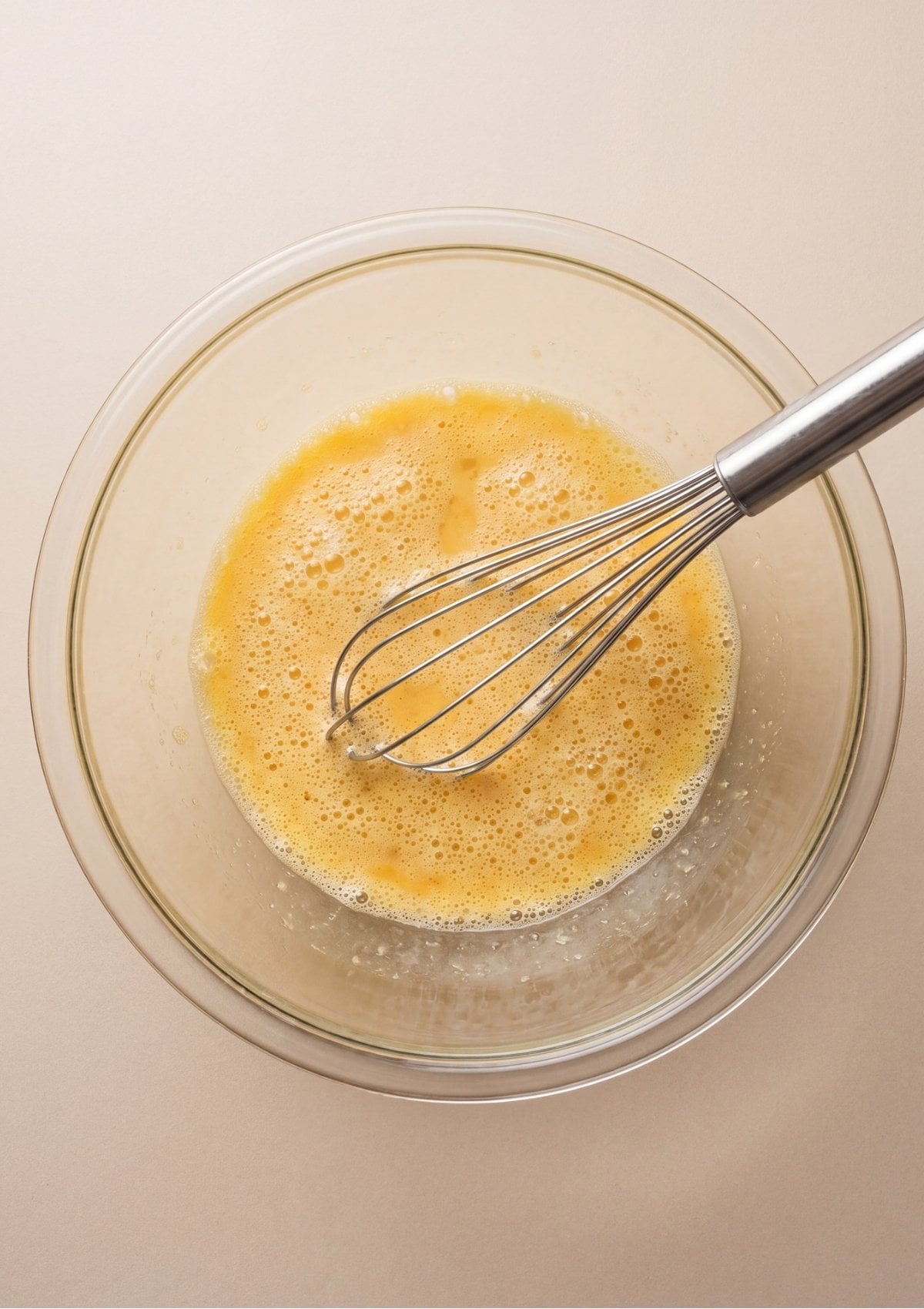 A stainless steel whisk mixing whisked eggs, milk, and melted butter into a bubbly frothy liquid inside a clear glass bowl