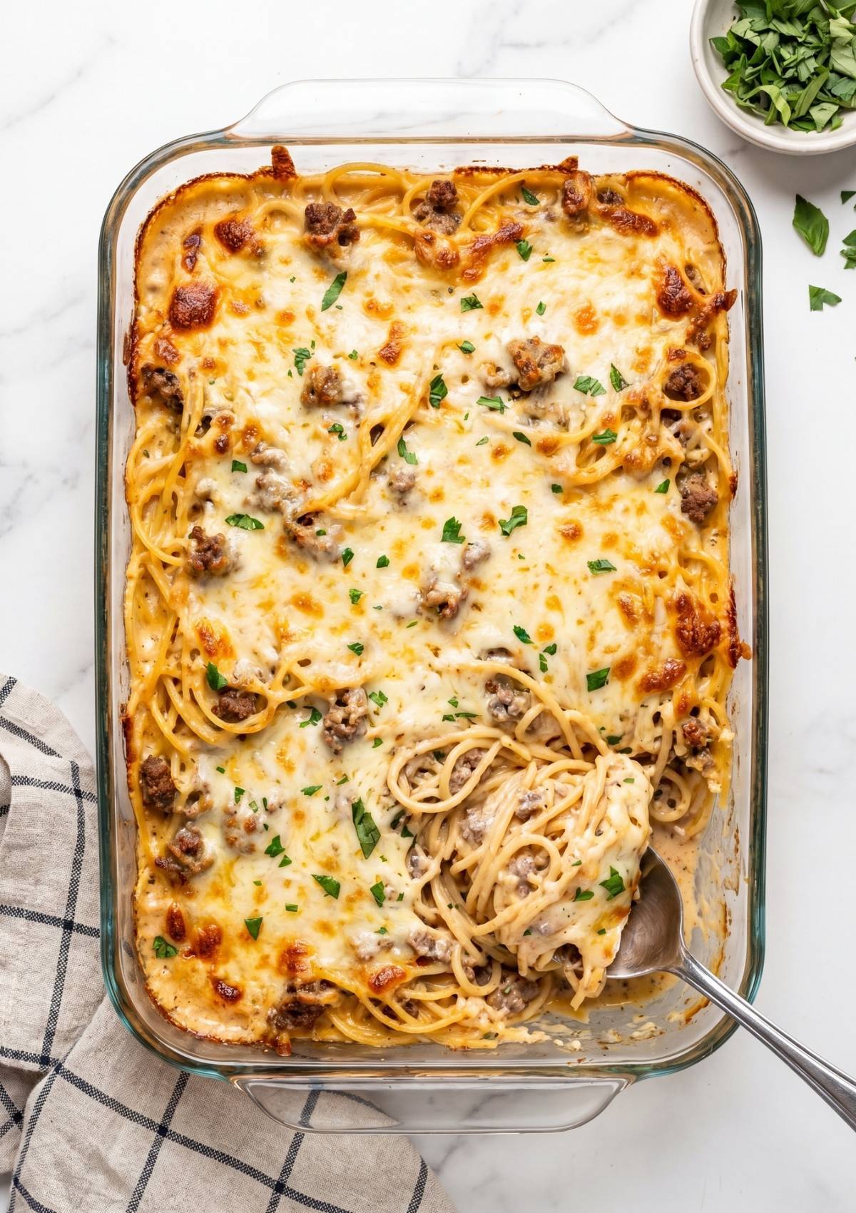 A large glass casserole dish of golden-brown baked Cowboy Spaghetti with Cream Cheese, with a metal spoon scooping out a serving of cheesy pasta and meat.