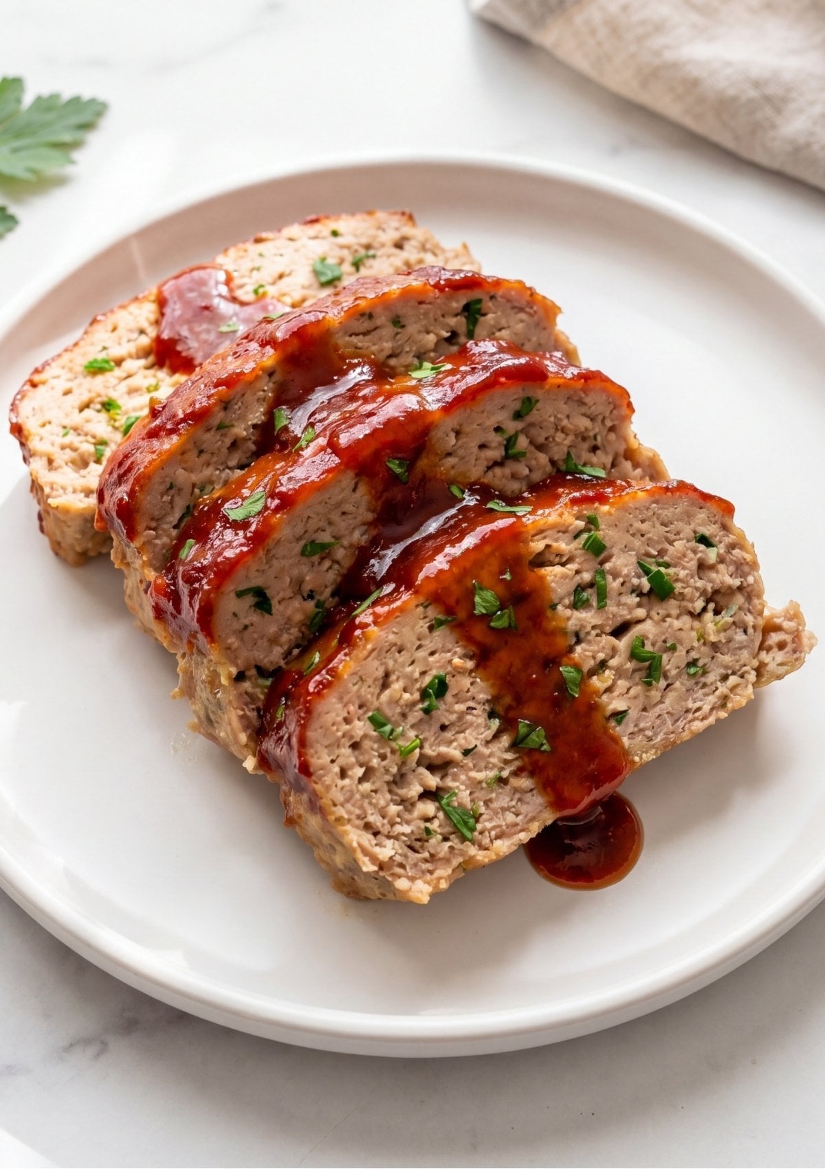 Close up of several thick slices of ground turkey meatloaf with a glossy red glaze and chopped green parsley on a white round plate.
