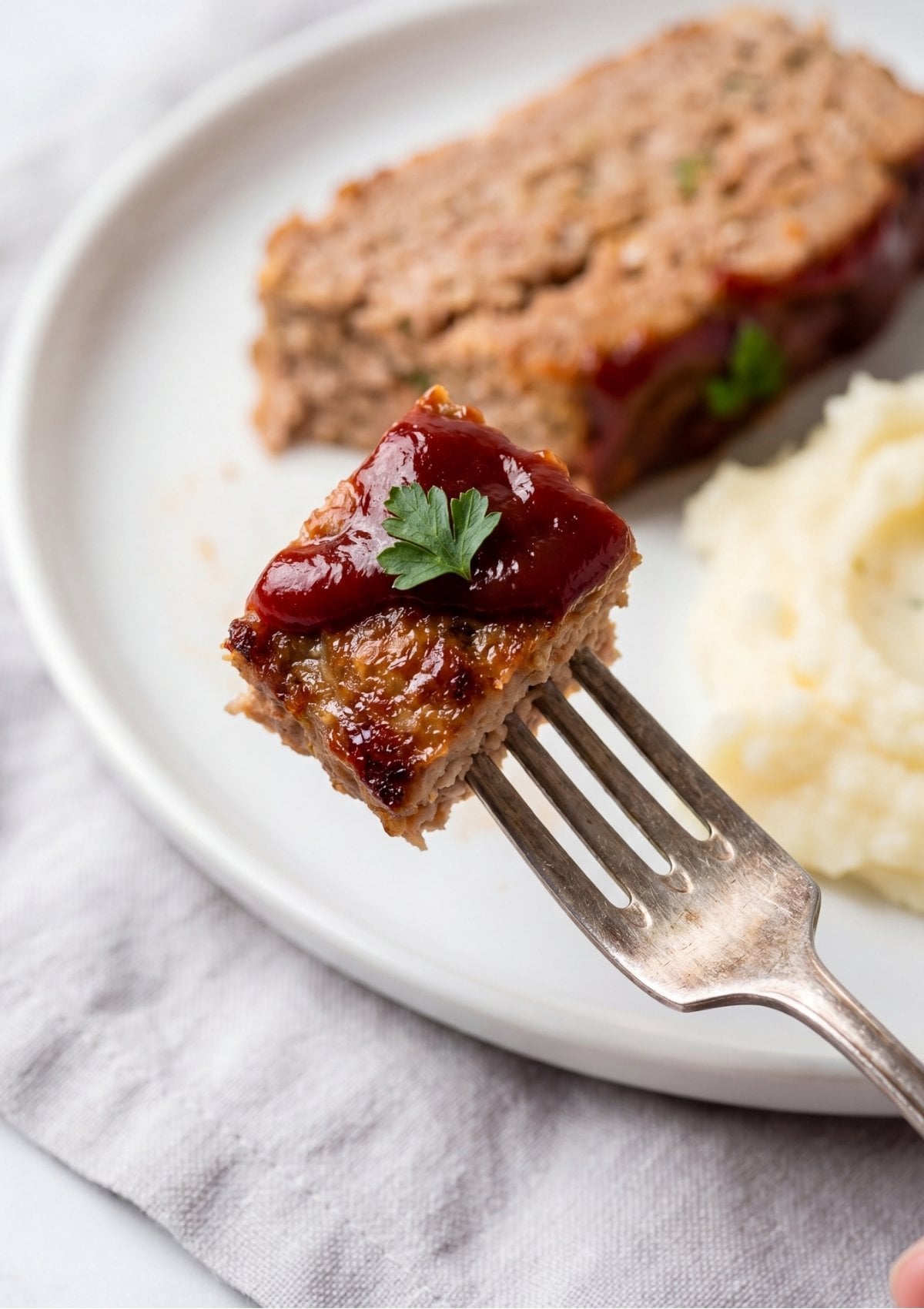 A single bite-sized piece of glazed turkey meatloaf garnished with a parsley leaf on the prongs of a silver fork, with a plate of mashed potatoes and a meatloaf slice blurred in the background.
