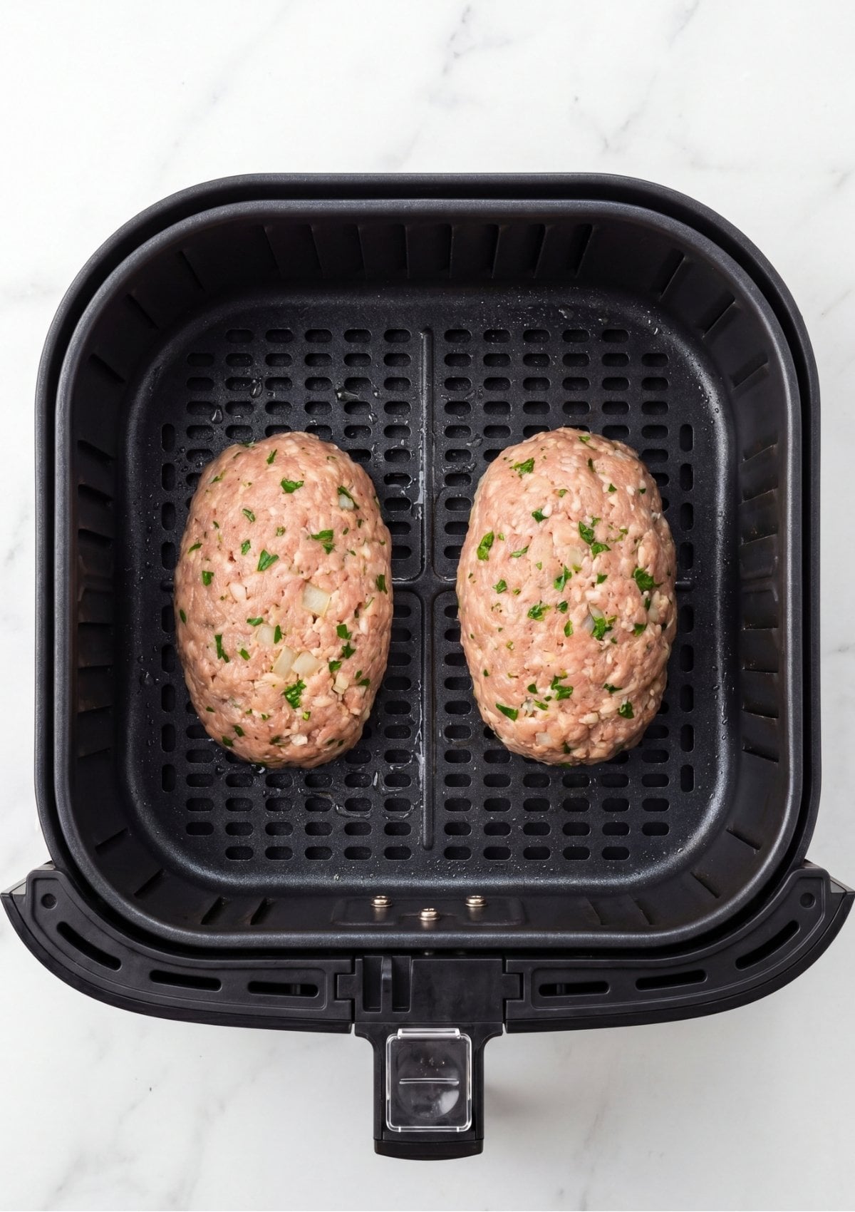 Two raw, oval-shaped ground turkey meatloaves mixed with visible flecks of green parsley and onion sitting inside a black air fryer basket before cooking